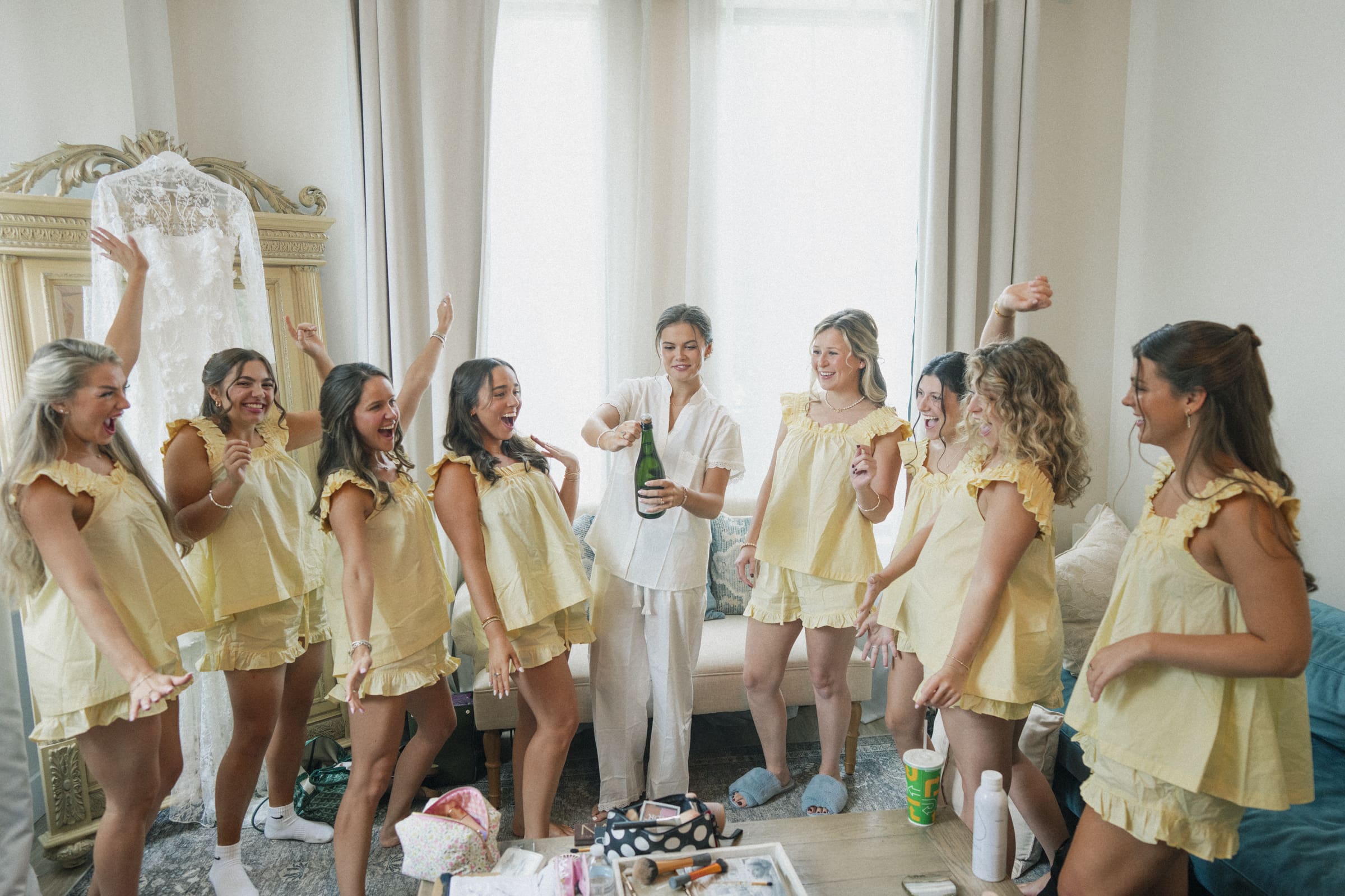 A bride in white pajamas pops a champagne bottle, surrounded by eight bridesmaids in matching yellow pajamas, all cheering and celebrating together in a bright room. A wedding dress hangs in the background.
