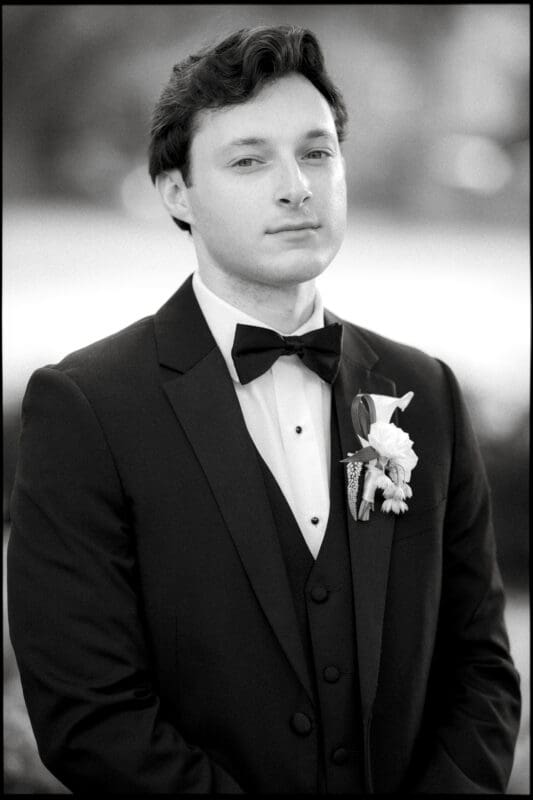 A man in a black tuxedo with a bow tie and boutonnière stands facing the camera, looking serious. The background is softly blurred, giving the portrait a formal and elegant feel.
