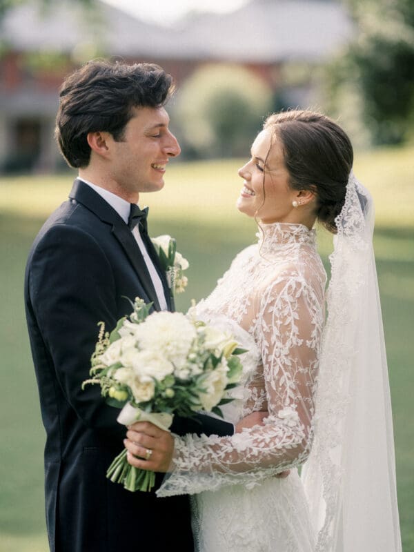 A bride in a white lace dress and veil smiles at a groom in a dark suit and bow tie. She holds a bouquet of white flowers as they stand outdoors on a sunny day, gazing happily at each other.
