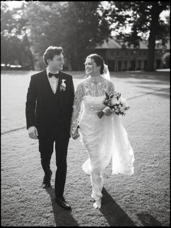 A bride and groom walk hand in hand on a grassy lawn, smiling at each other. The bride wears a long lace dress and veil, holding a bouquet, while the groom is in a black tuxedo. Sunlight creates a soft, romantic atmosphere.