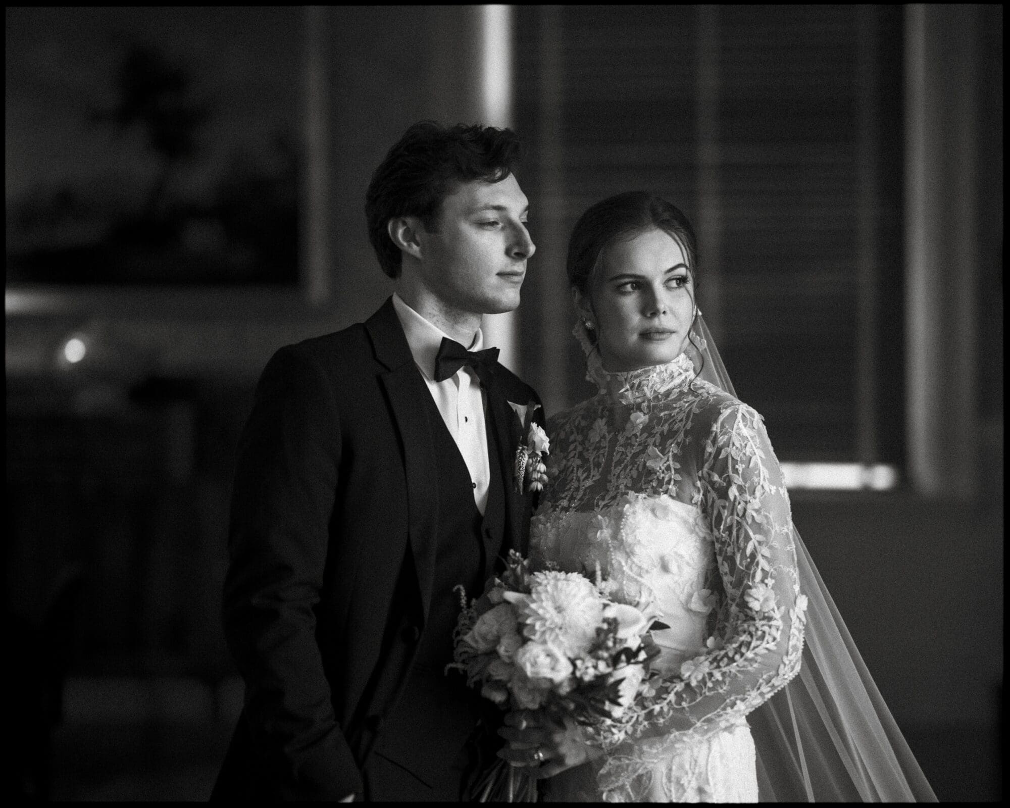 A bride in a lace dress and veil stands beside a groom in a tuxedo, both looking thoughtfully into the distance. The bride holds a bouquet of flowers. The image is in black and white.
