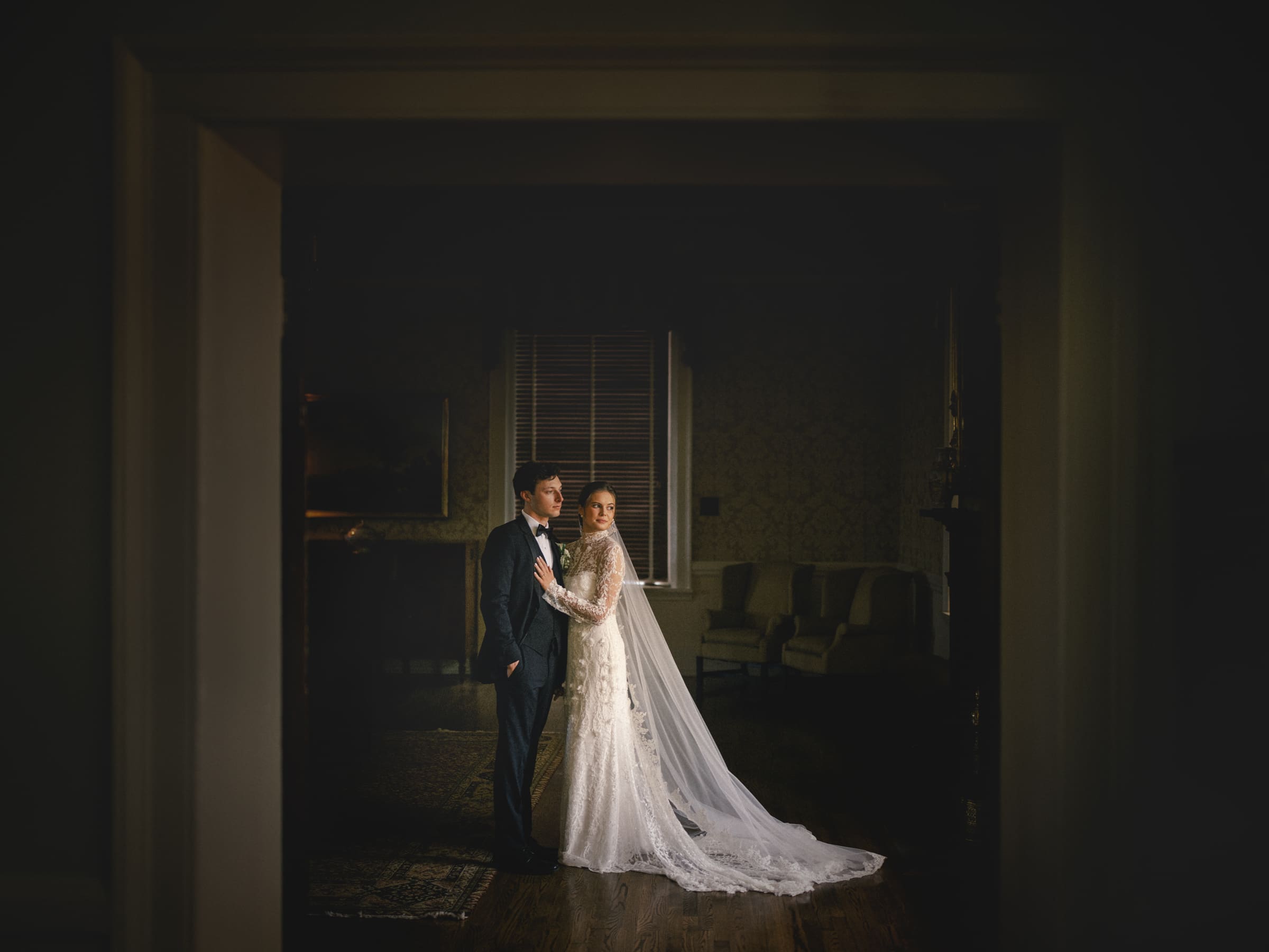 A bride and groom stand together in a dimly lit room, framed by a doorway. The bride wears a long white gown with a train, and the groom is in a dark suit. Soft light highlights them against a dark, elegant background.