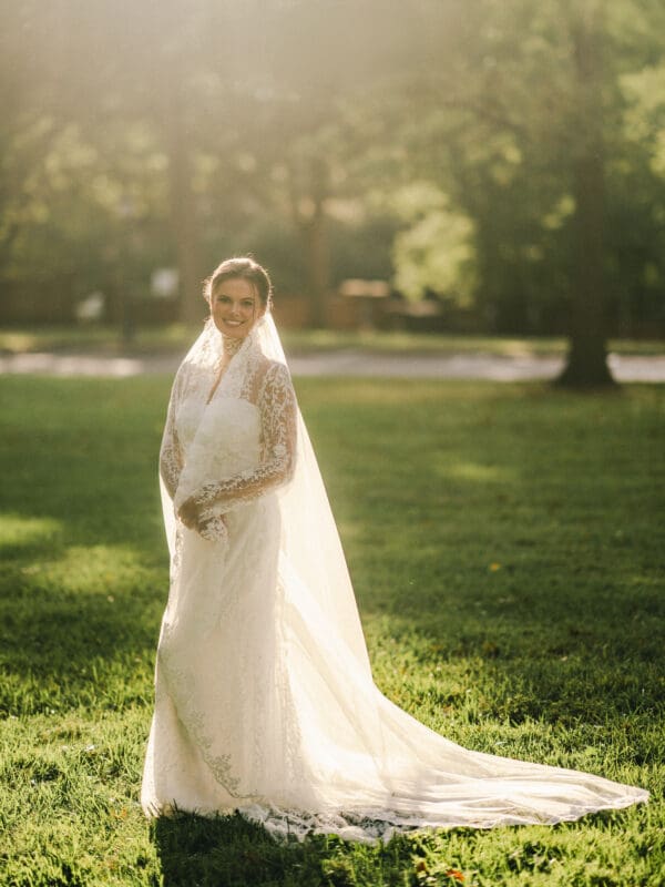 A bride in a lace wedding dress and long veil stands on green grass, smiling in soft, golden sunlight with trees and a blurred background behind her.