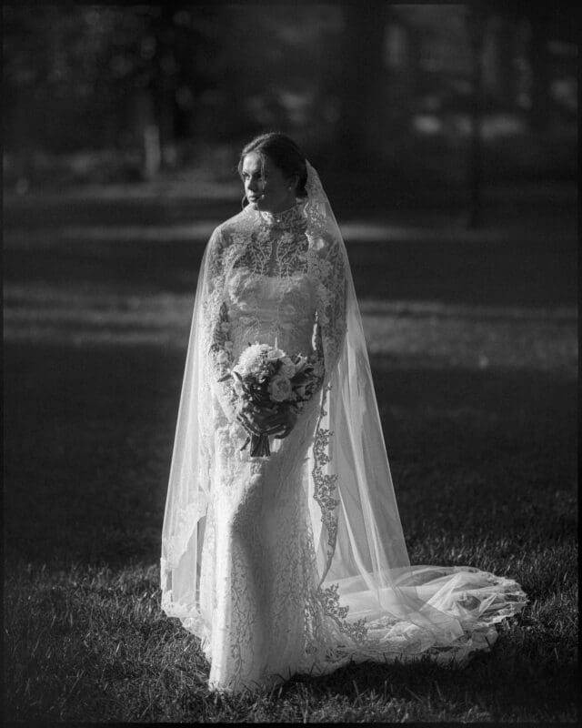 A bride in a long lace wedding dress and veil stands outdoors on grass, holding a bouquet of flowers, looking to the side. Sunlight softly illuminates her and creates a gentle, elegant atmosphere.