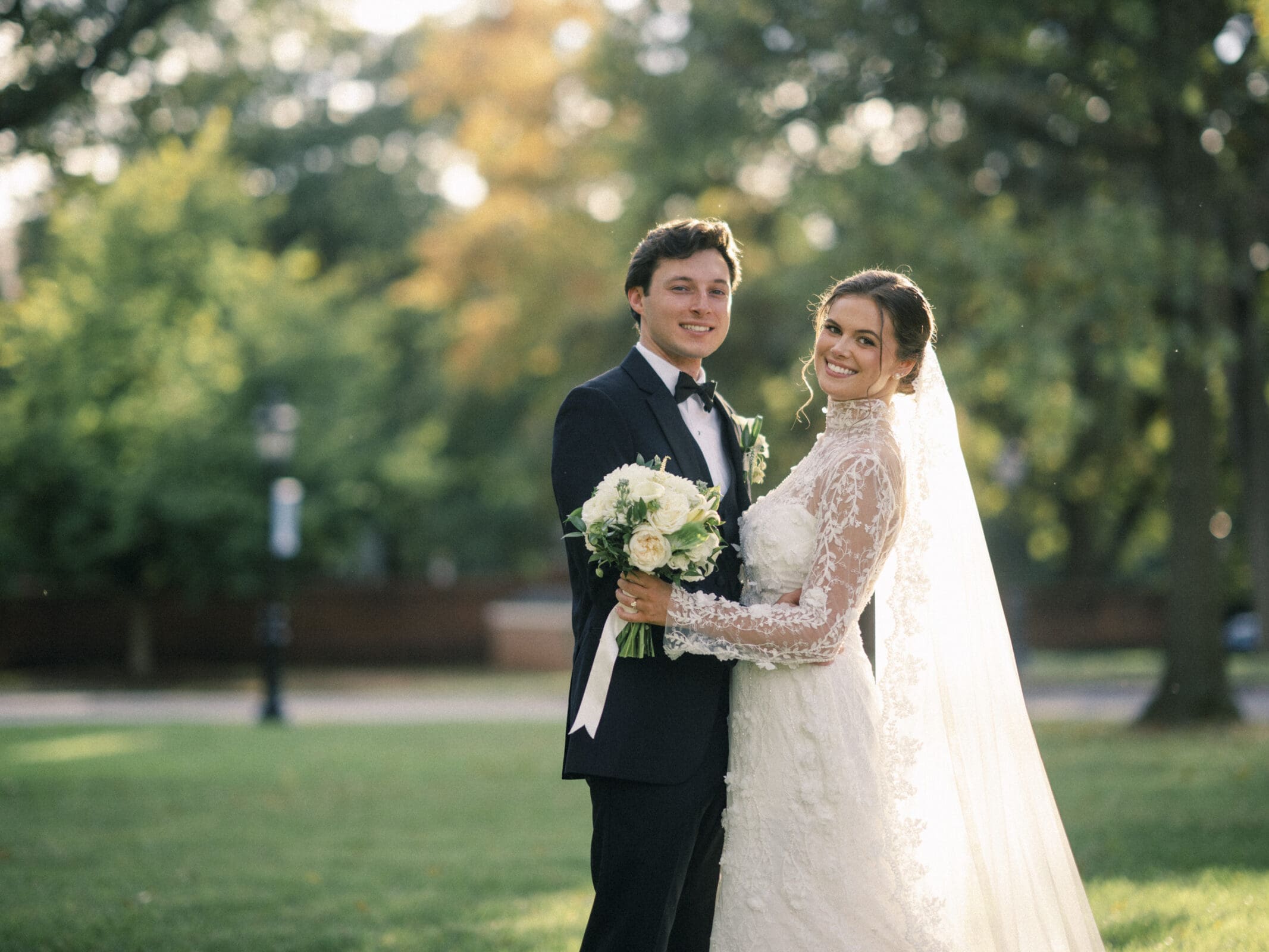A bride in a lace wedding dress and veil stands smiling with a groom in a tuxedo, holding a bouquet, outdoors in a sunlit park with green trees in the background.
