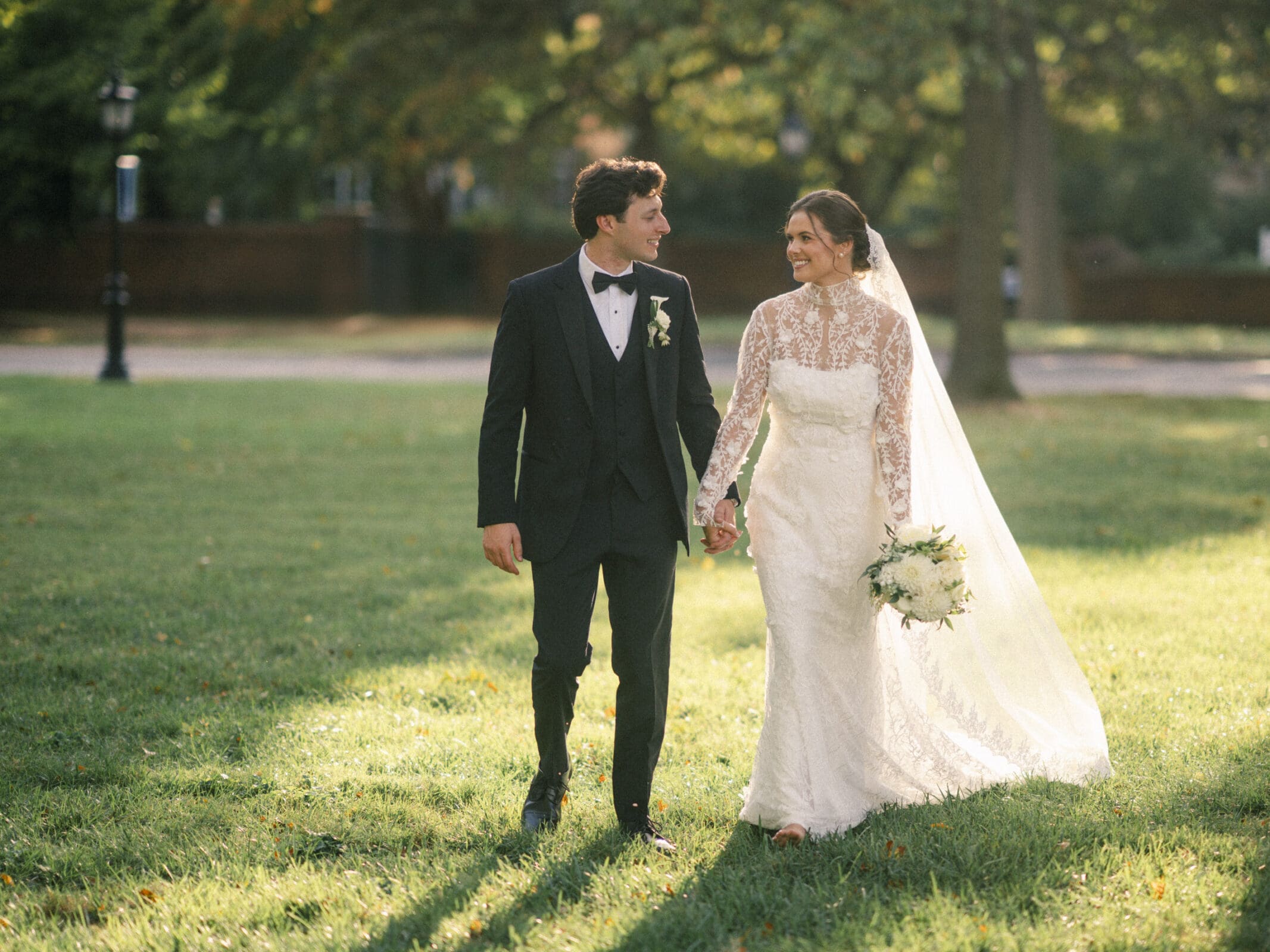 A bride and groom walk hand in hand on a grassy lawn. The groom wears a black tuxedo, and the bride wears a long-sleeved lace wedding dress with a veil, holding a bouquet. Sunlight filters through trees in the background.