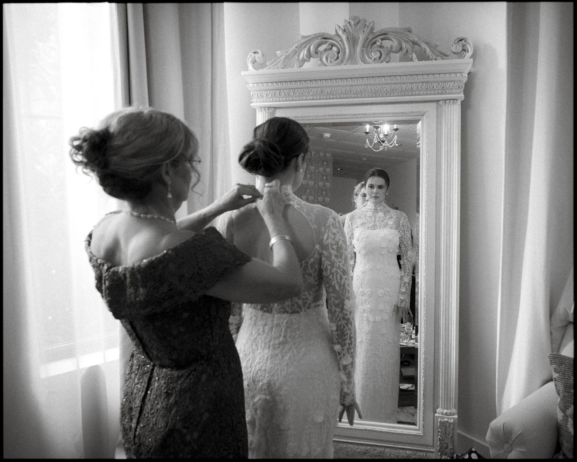 A woman helps a bride fasten her lace wedding dress in front of an ornate mirror. The bride’s reflection shows her looking ahead, while the woman stands behind adjusting the dress in a softly lit room.