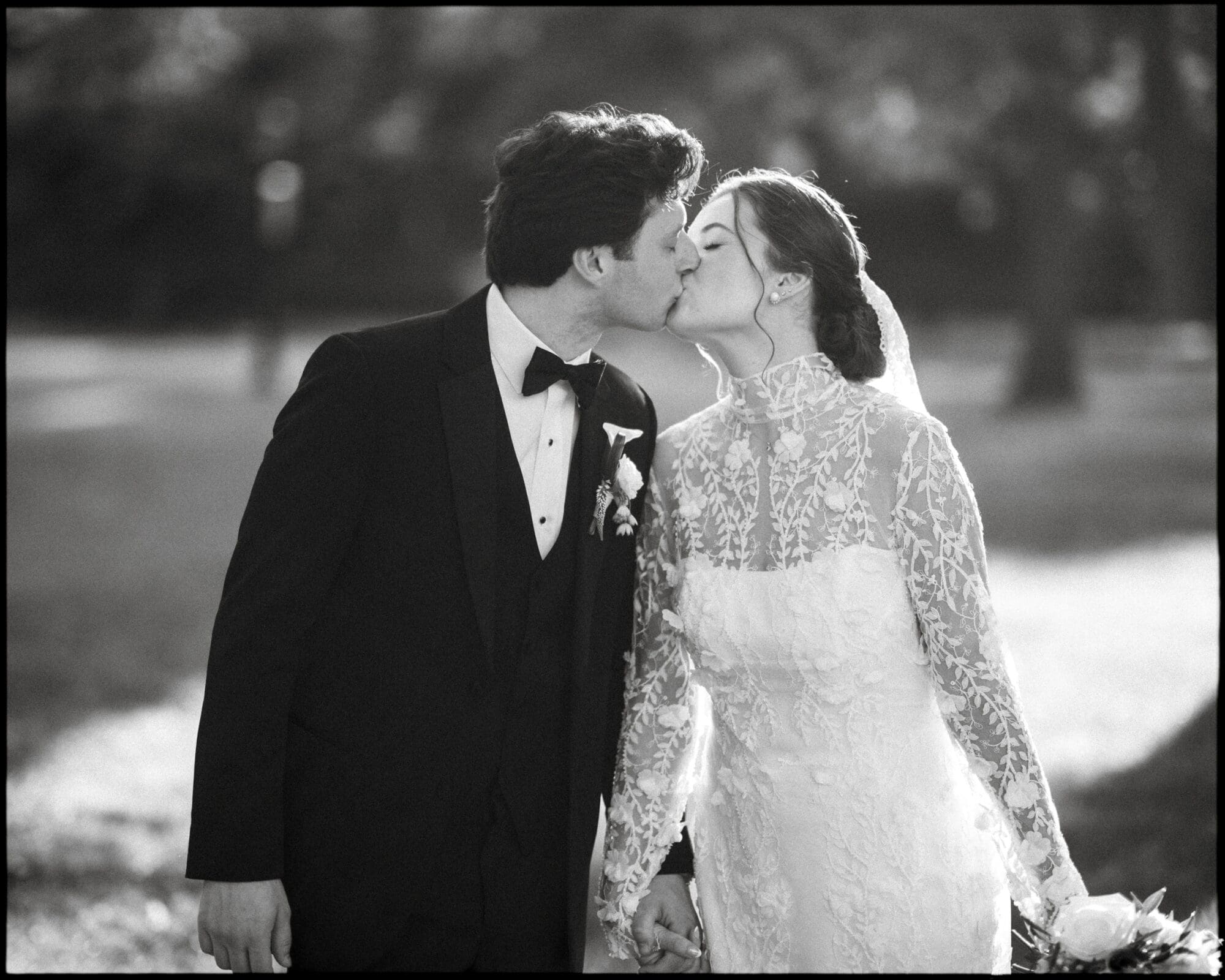 A bride and groom kiss outdoors, holding hands. The groom wears a black tuxedo with a bow tie, and the bride wears a long-sleeved lace wedding dress and veil. Sunlight softly illuminates the scene.