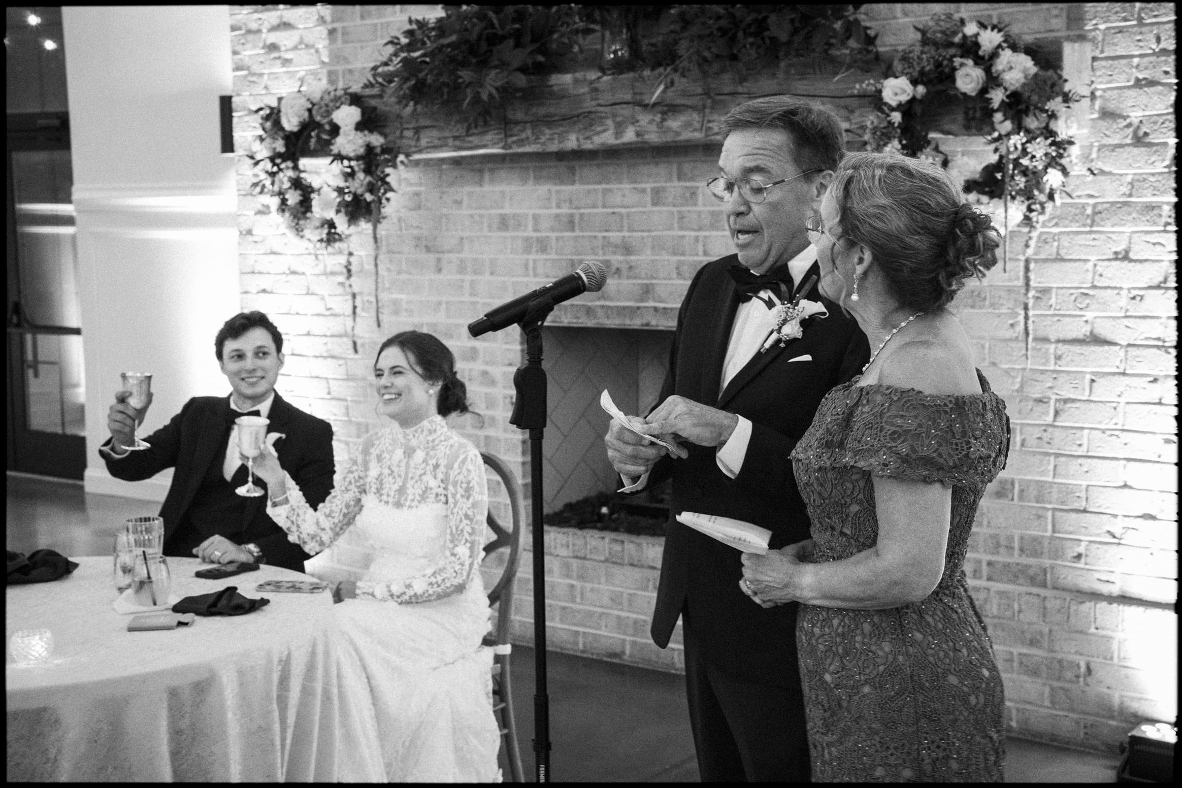 A couple stands at a microphone giving a speech at a wedding reception, while the bride and groom, seated at a table, smile and raise their glasses in a toast. The setting features a decorated fireplace with flowers.