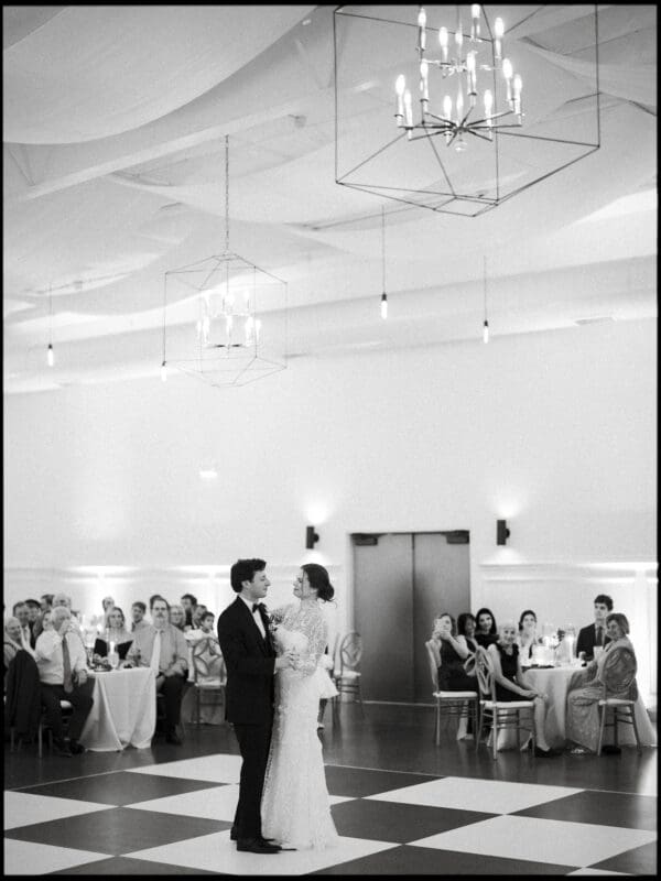 A bride and groom share their first dance on a checkered floor in a bright, elegant reception hall, surrounded by seated guests watching them. Chandeliers hang from the ceiling above.