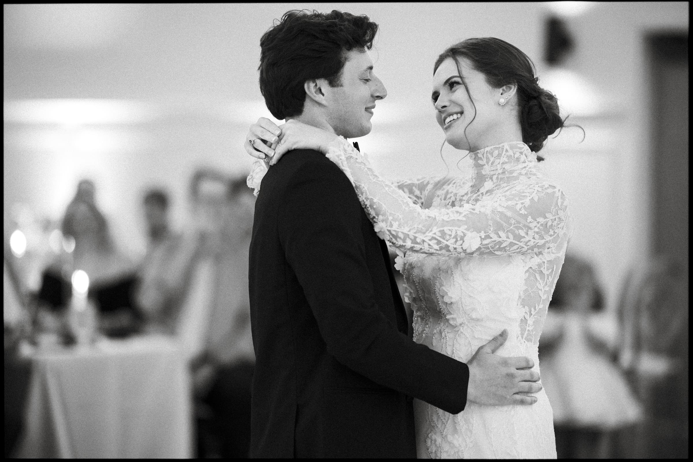 A bride and groom smile at each other while dancing closely at their wedding reception. The bride wears a lace dress, and the groom is in a suit. Guests are seated and watching in the softly lit background.