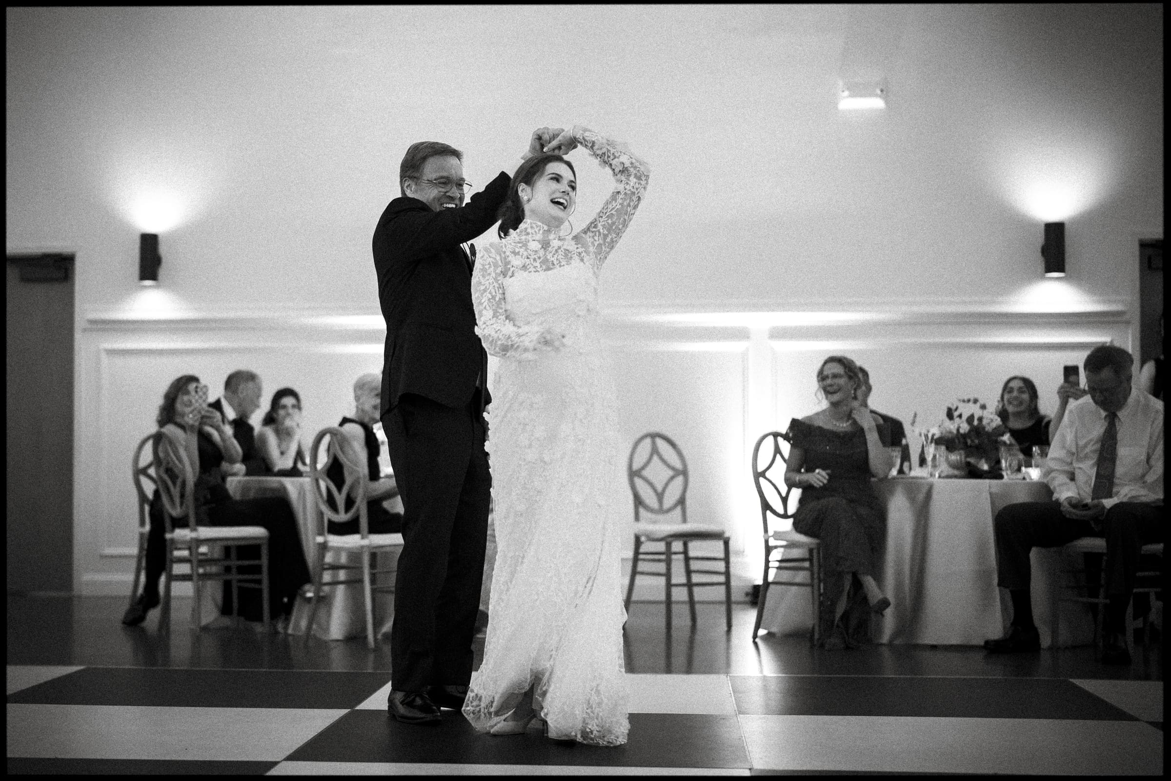 A smiling bride in a long-sleeved lace gown dances with an older man in a suit, likely her father, on a checkered dance floor. Seated guests in formal attire watch and smile in the background.
