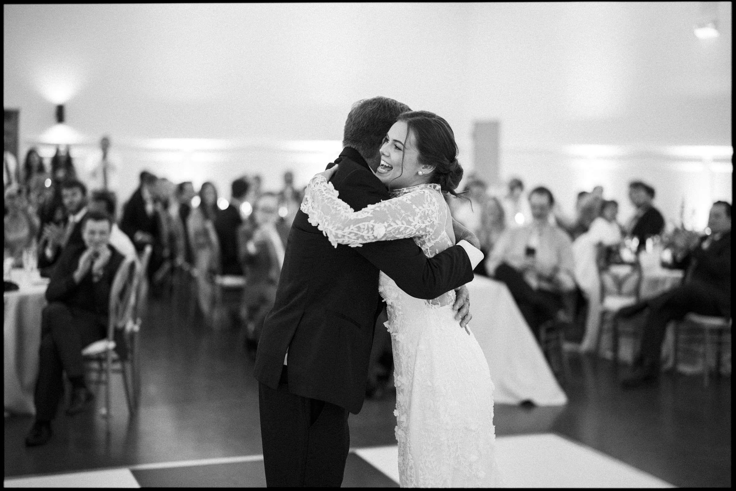 A bride in a lace wedding dress and a man in a suit share a joyful hug on the dance floor, surrounded by seated guests at a wedding reception.