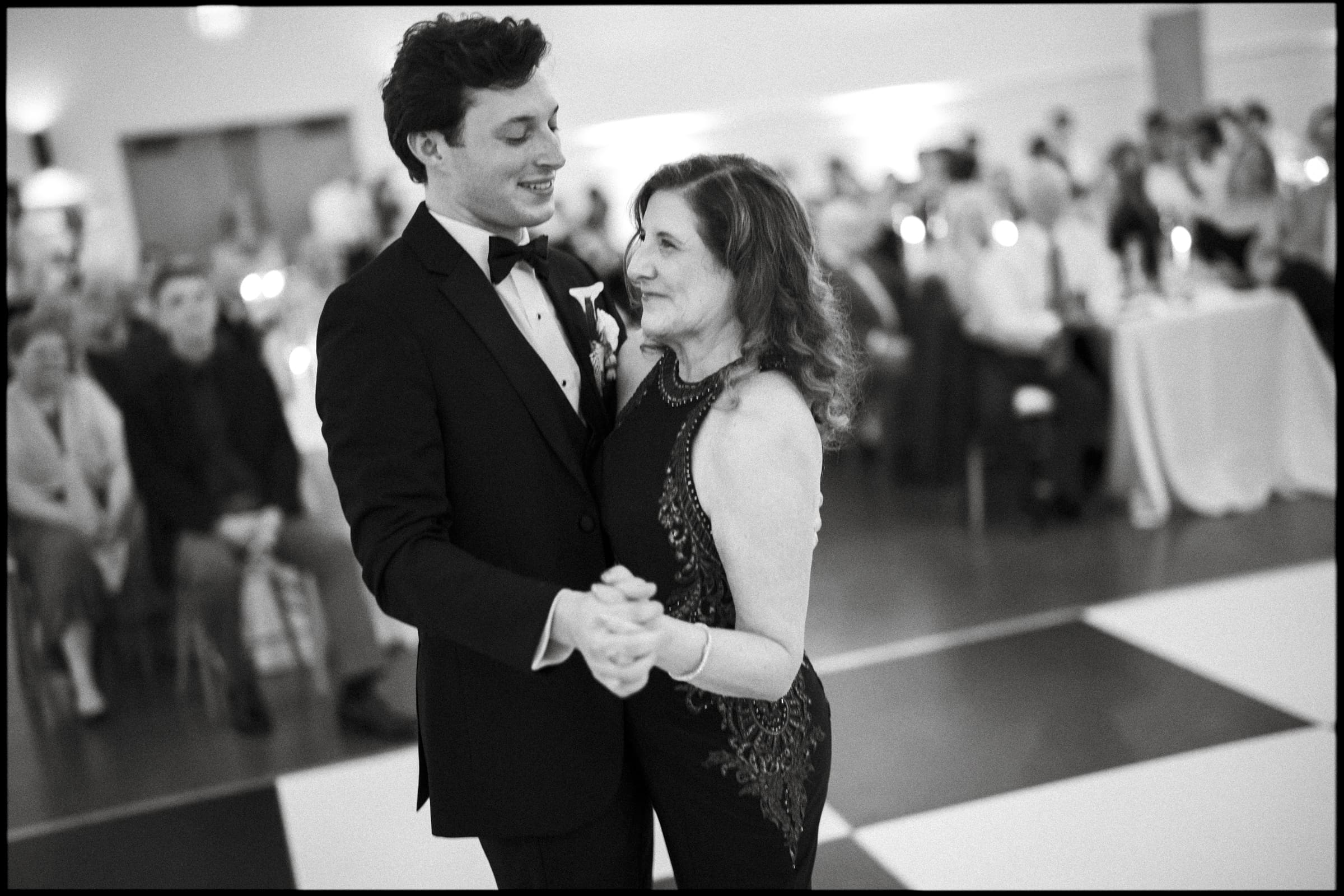 A young man in a tuxedo and a woman in a formal dress smile at each other while dancing together on a black-and-white checkered dance floor, with people seated and watching in the background.