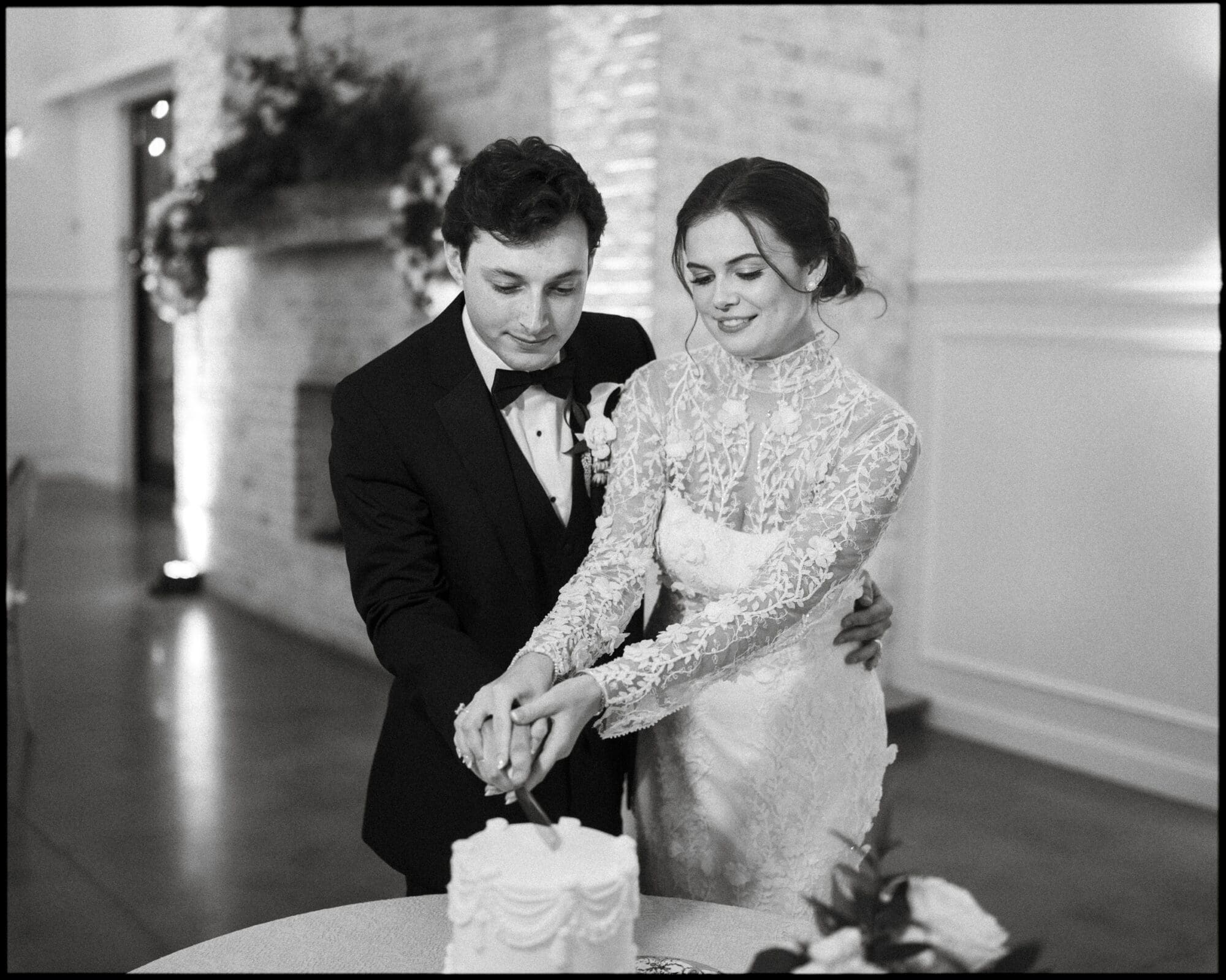 A bride and groom in formal attire smile as they cut a small wedding cake together in an elegant, softly lit venue with floral decorations.
