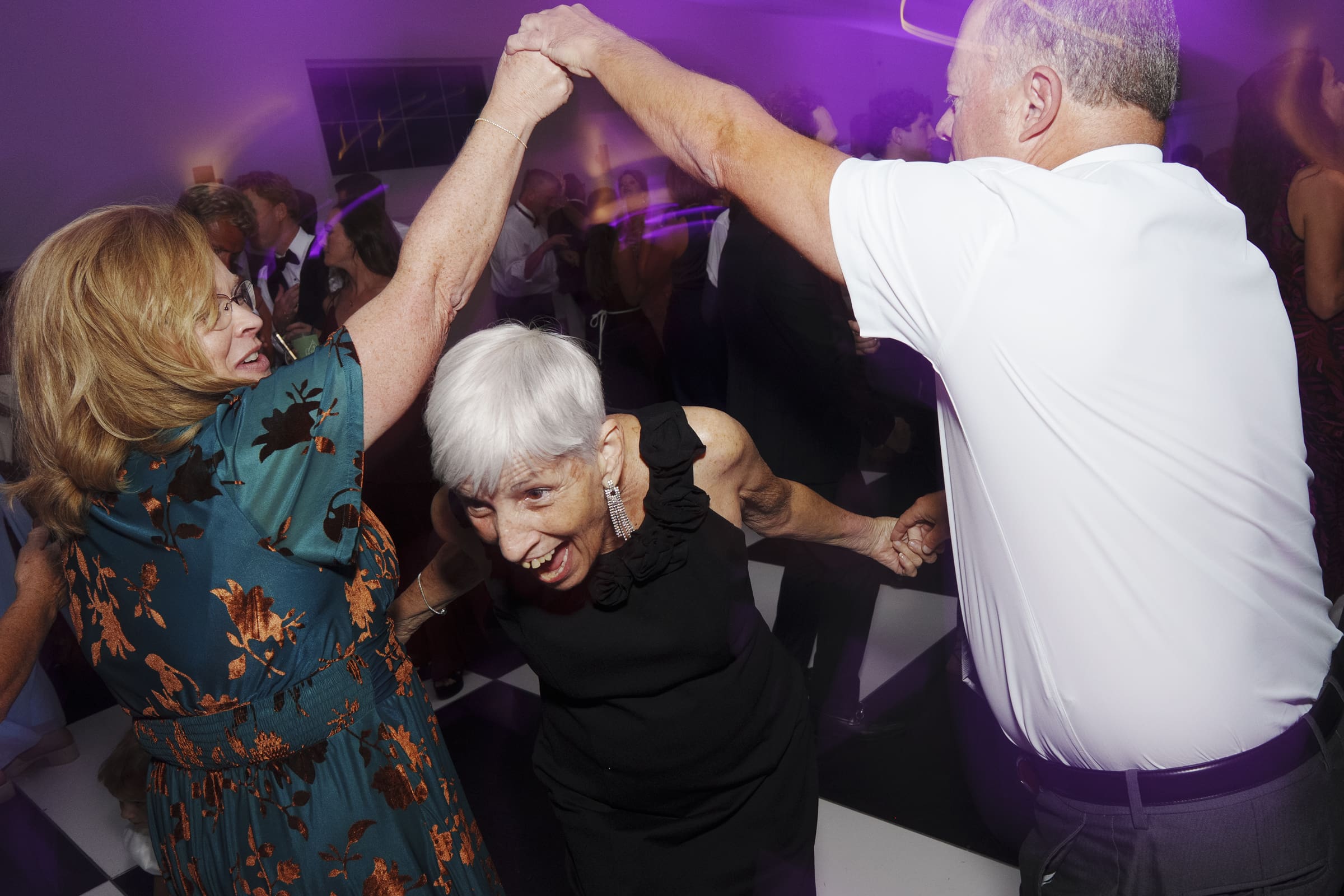 Three older adults dance joyfully together at a lively indoor event. One woman in a floral dress and another in black are spinning under a mans raised arm. The scene is vibrant with purple lighting and a joyful atmosphere.
