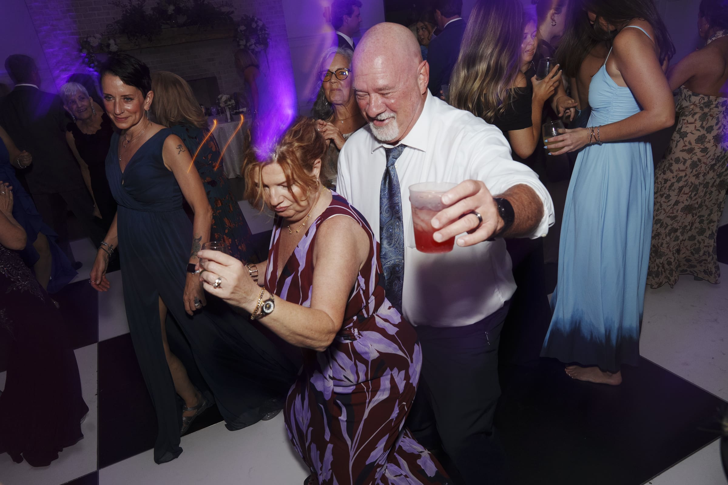 A group of people dance energetically at a party, drinks in hand, under purple lighting. Smiling and laughing, they wear formal attire, and the mood is joyful and celebratory.