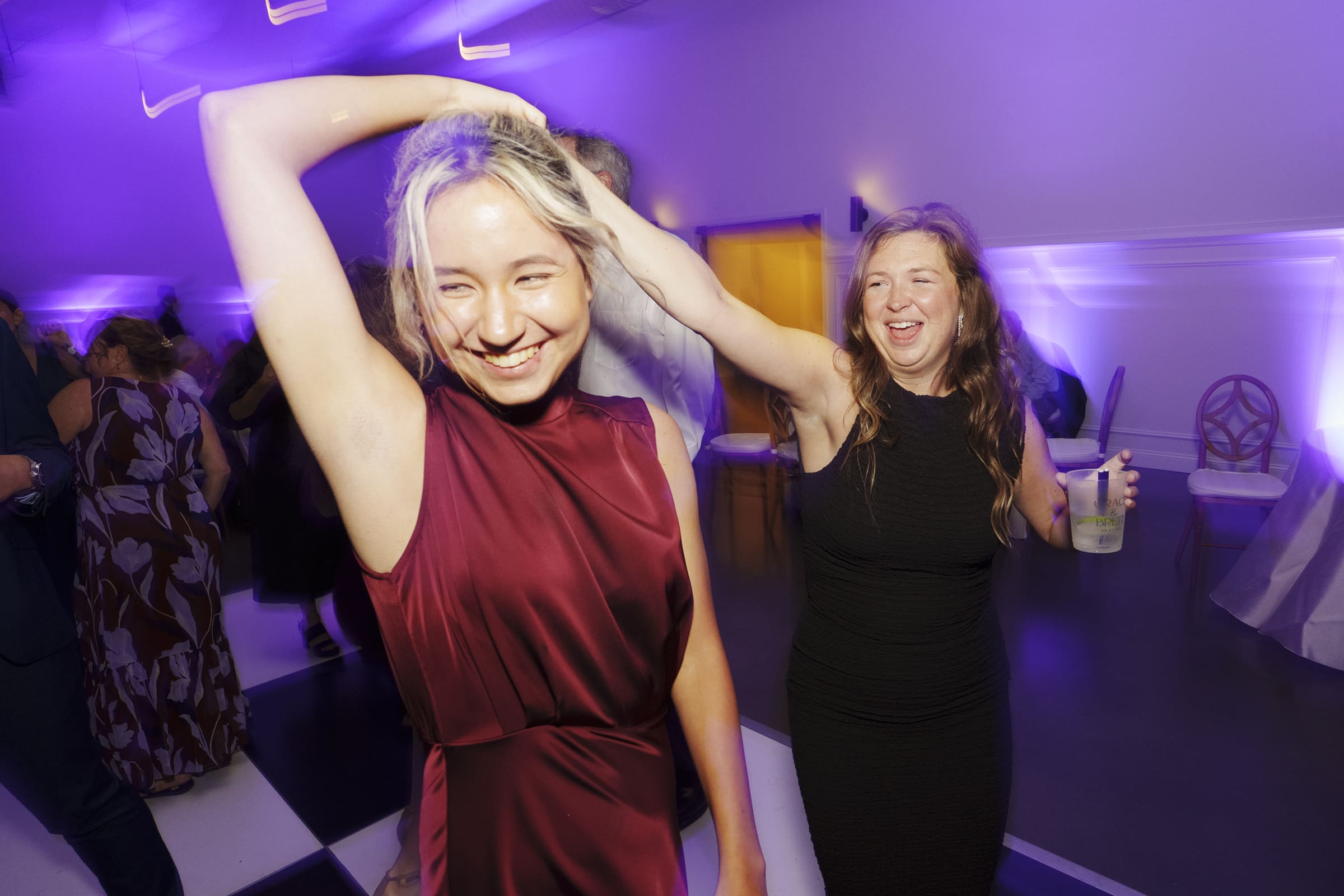 Two women are dancing and laughing together in a room with purple lighting. One wears a sleeveless burgundy dress and raises her arm, while the other, in a black dress, holds a drink and smiles brightly. Others are blurred in the background.