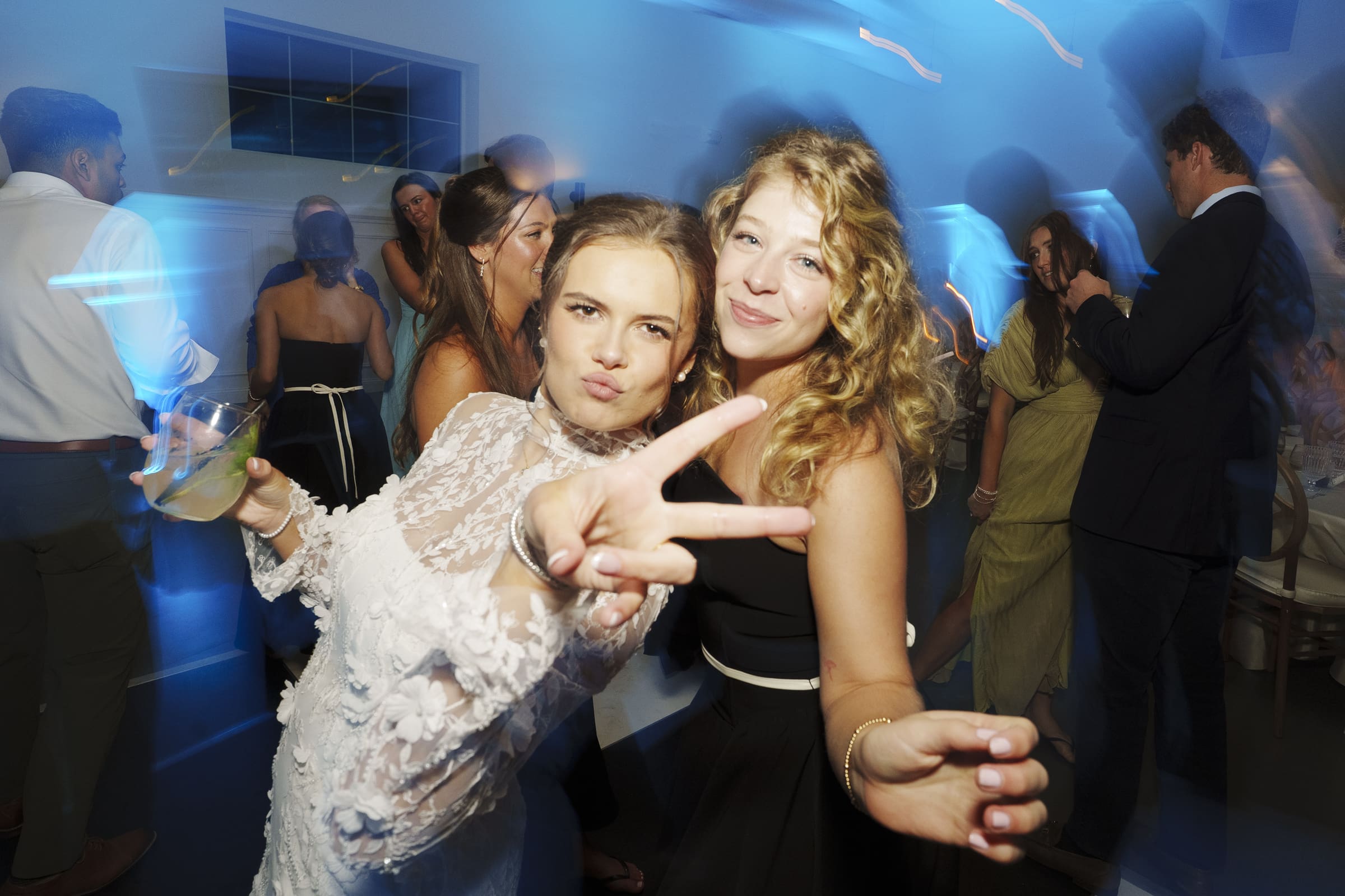 Two young women pose and smile at a lively party, one flashing a peace sign towards the camera. Other guests are mingling and dancing in the background under blue lighting.