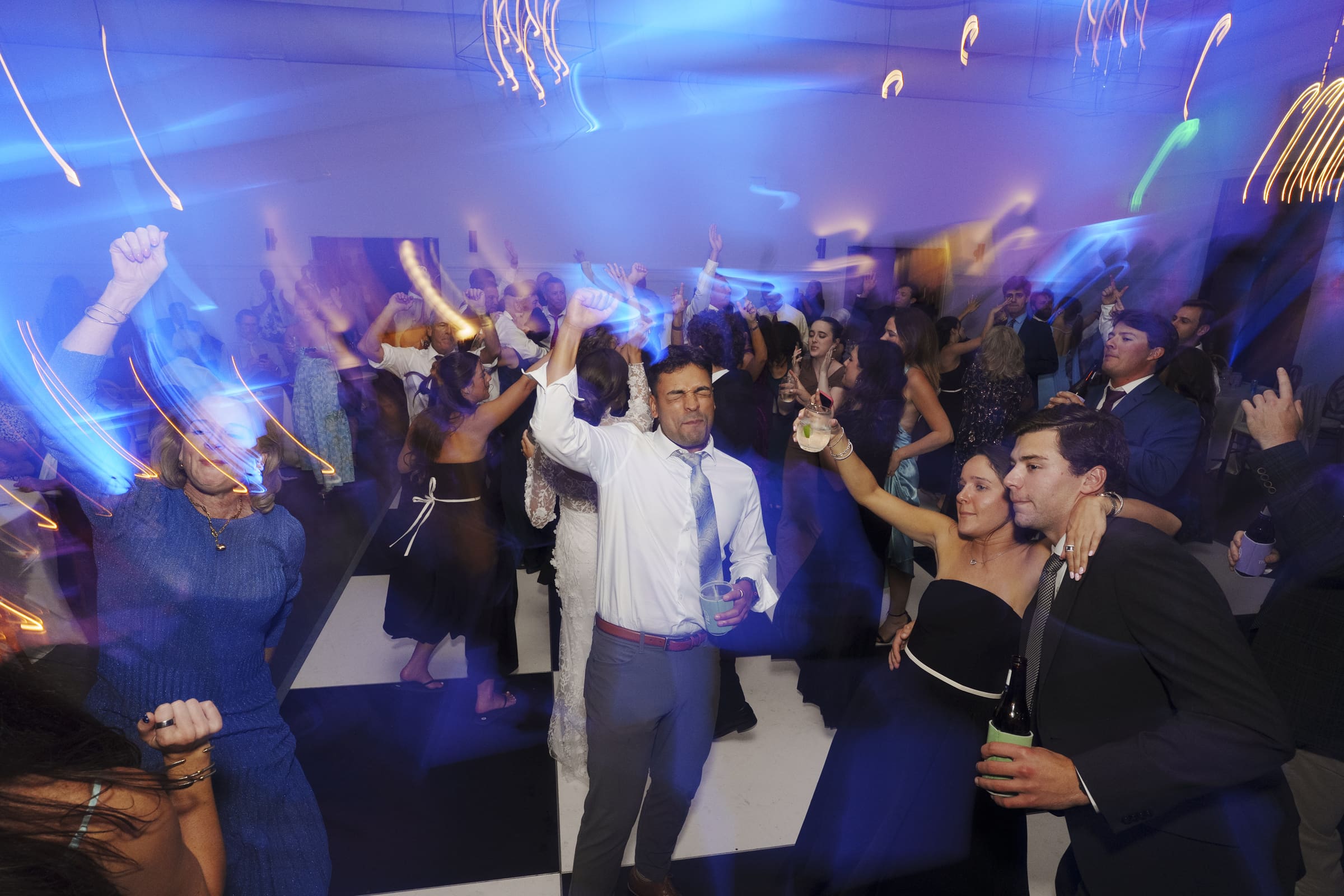 A lively group of people dance under colorful lights at a party. Some raise their arms and drinks, while others smile and move to the music. Blue and yellow light streaks add energy to the crowded, festive atmosphere.
