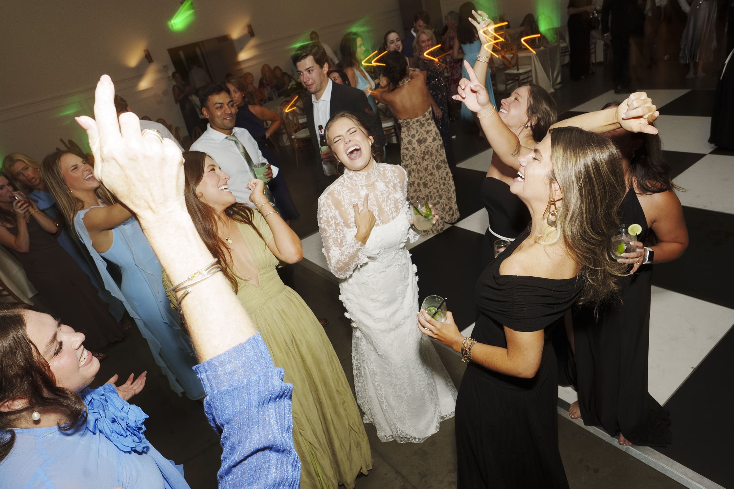 A joyful bride in a white lace gown laughs and dances with friends on a checkered dance floor at a lively wedding reception. Guests around her are smiling, raising their hands, and enjoying the celebration.