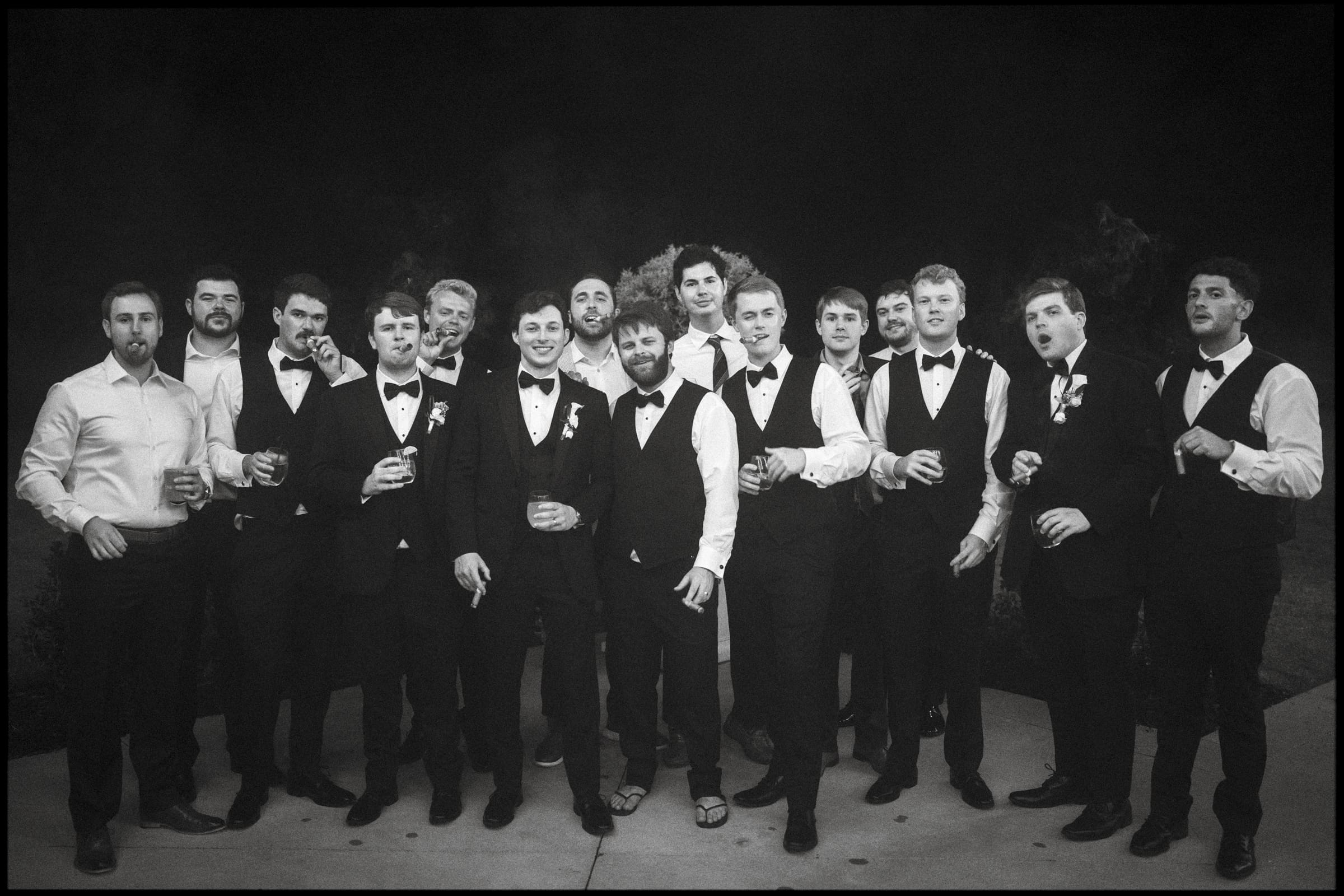 A group of men dressed in formal attire, mostly suits and bow ties, stand together outdoors at night. They are smiling and holding drinks, posing closely for a group photo. The image is black and white.