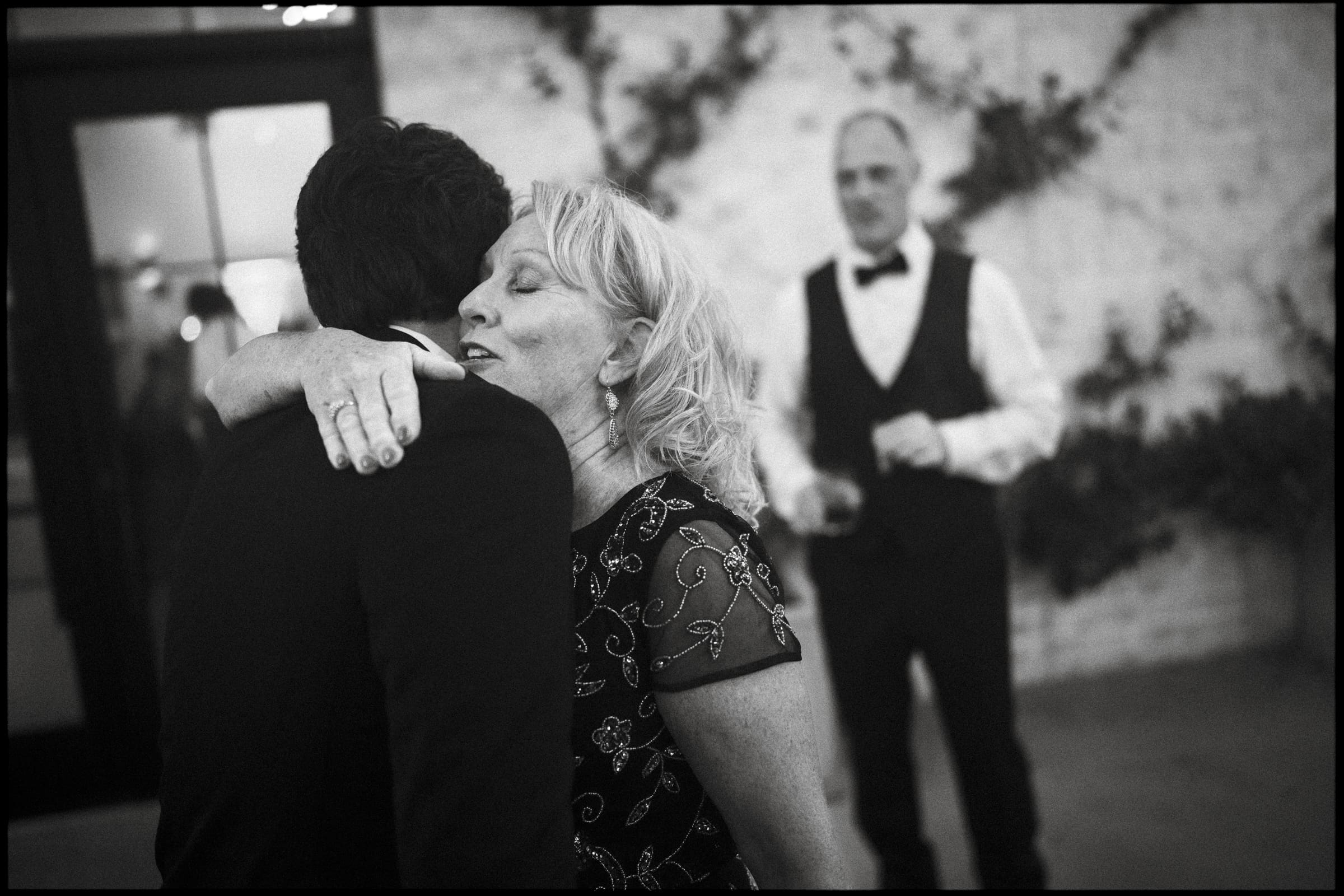 A woman with blonde hair hugs a man in a suit at an event, her eyes closed in a tender moment. In the background, a man in a bow tie and vest stands, watching them, with a textured wall behind him.