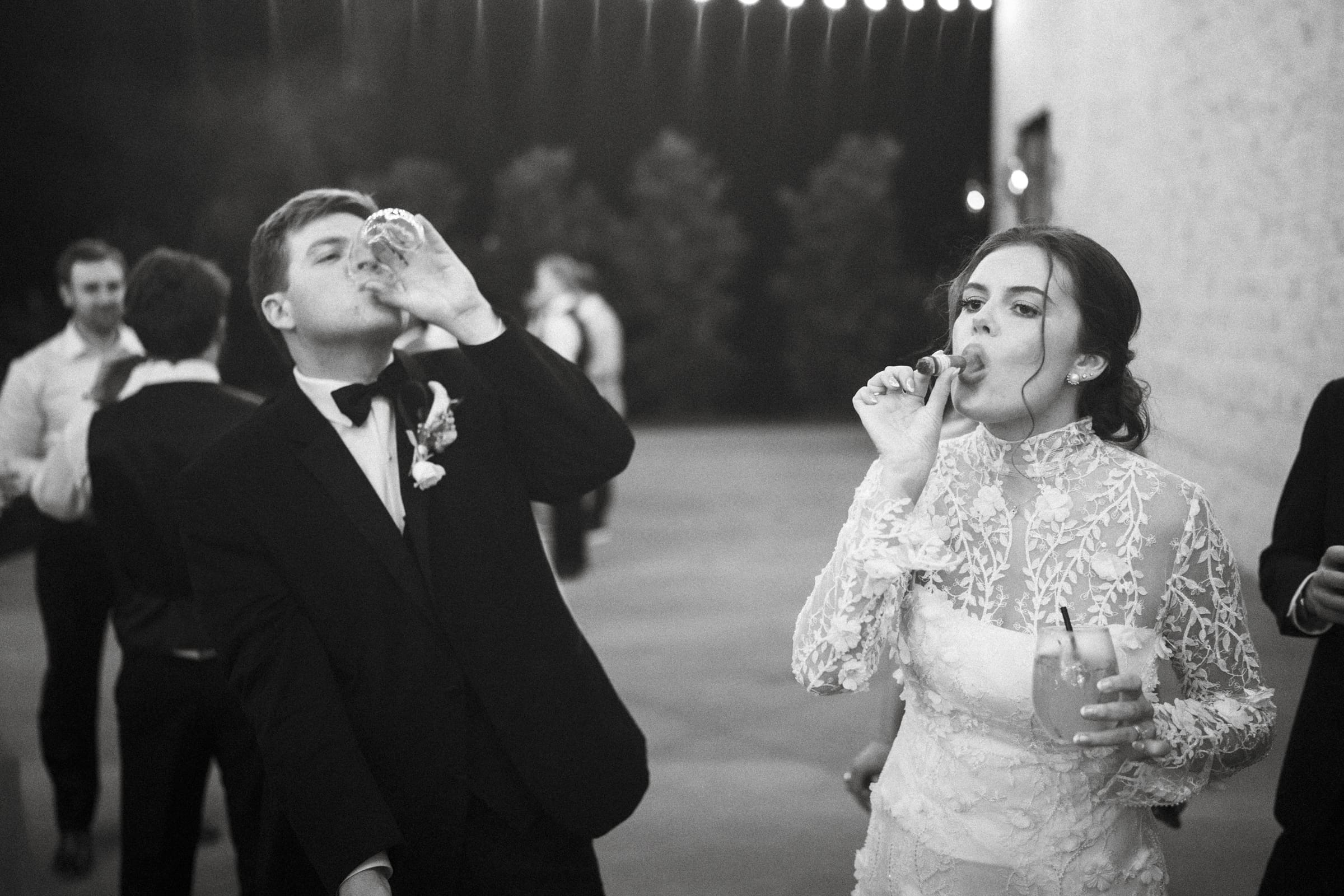 A bride in a lace dress and a groom in a suit are celebrating outdoors at night. The bride sips a drink through a straw, while the groom drinks from a glass. Other guests mingle in the blurred background.