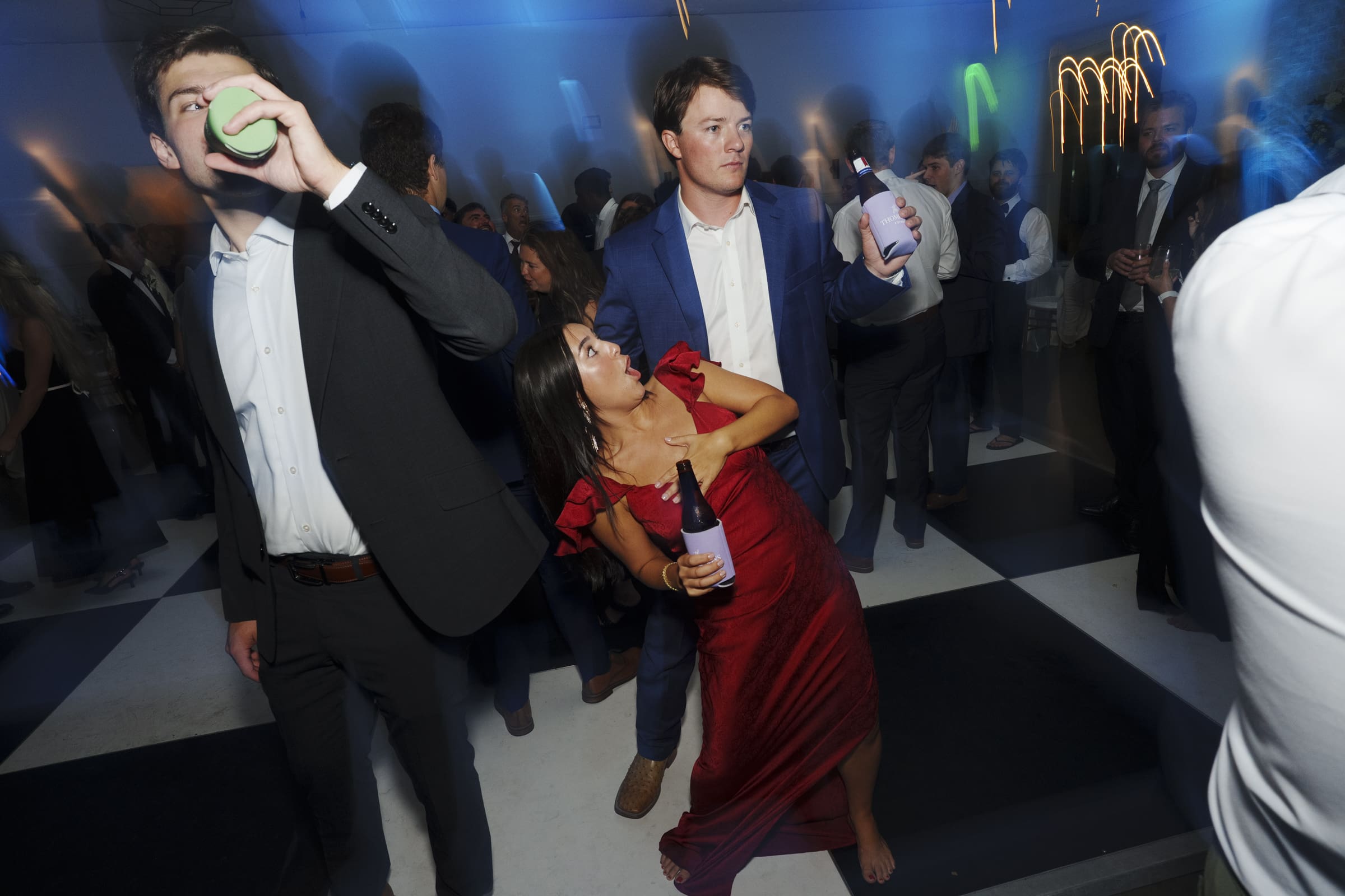 A barefoot woman in a red dress points and looks up while holding a drink, surrounded by men in suits at a lively party with colorful lights and a crowded dance floor.