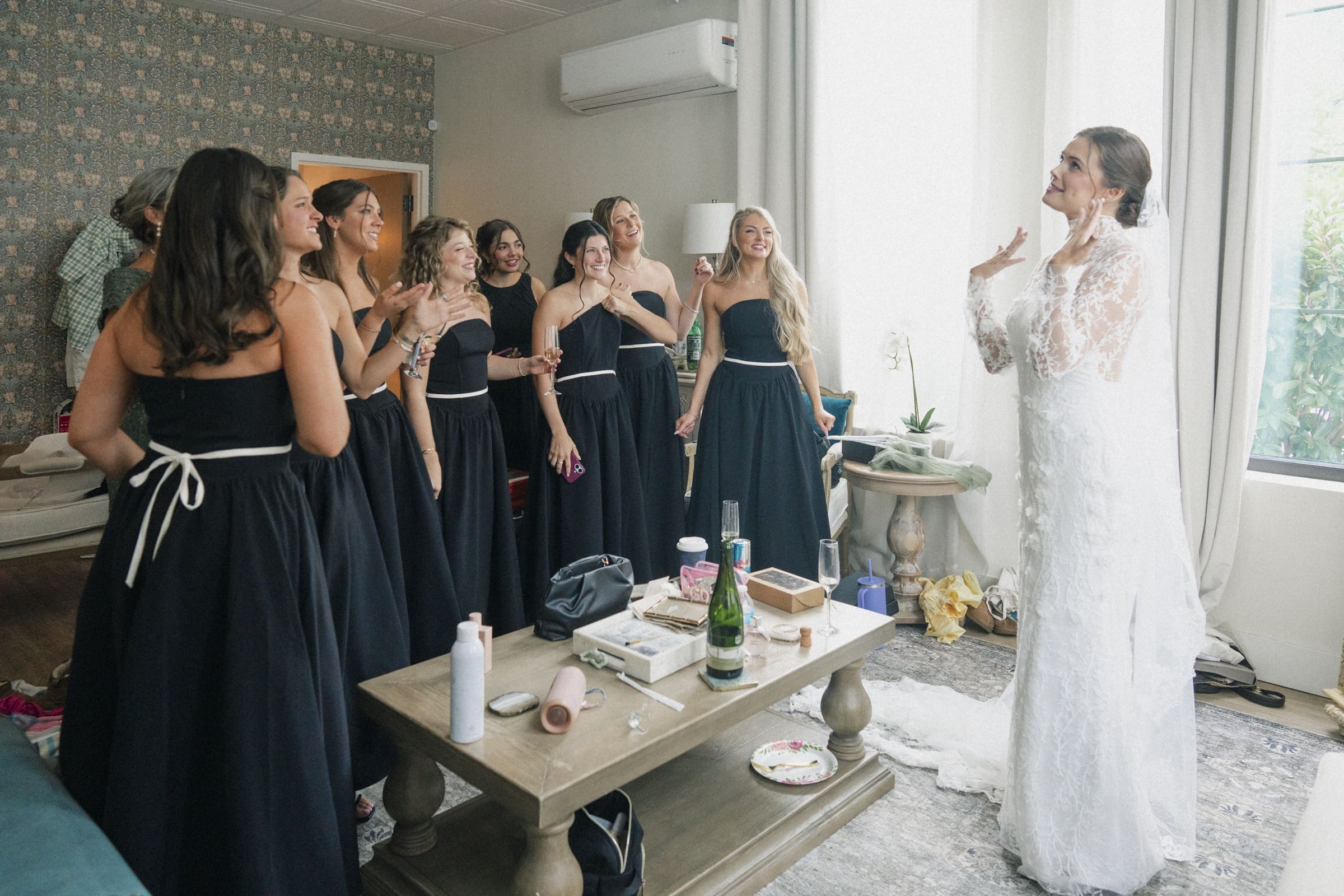 A bride in a white lace gown excitedly shows her dress to a group of bridesmaids in matching black dresses, who are smiling and reacting joyfully in a bright, softly lit room.