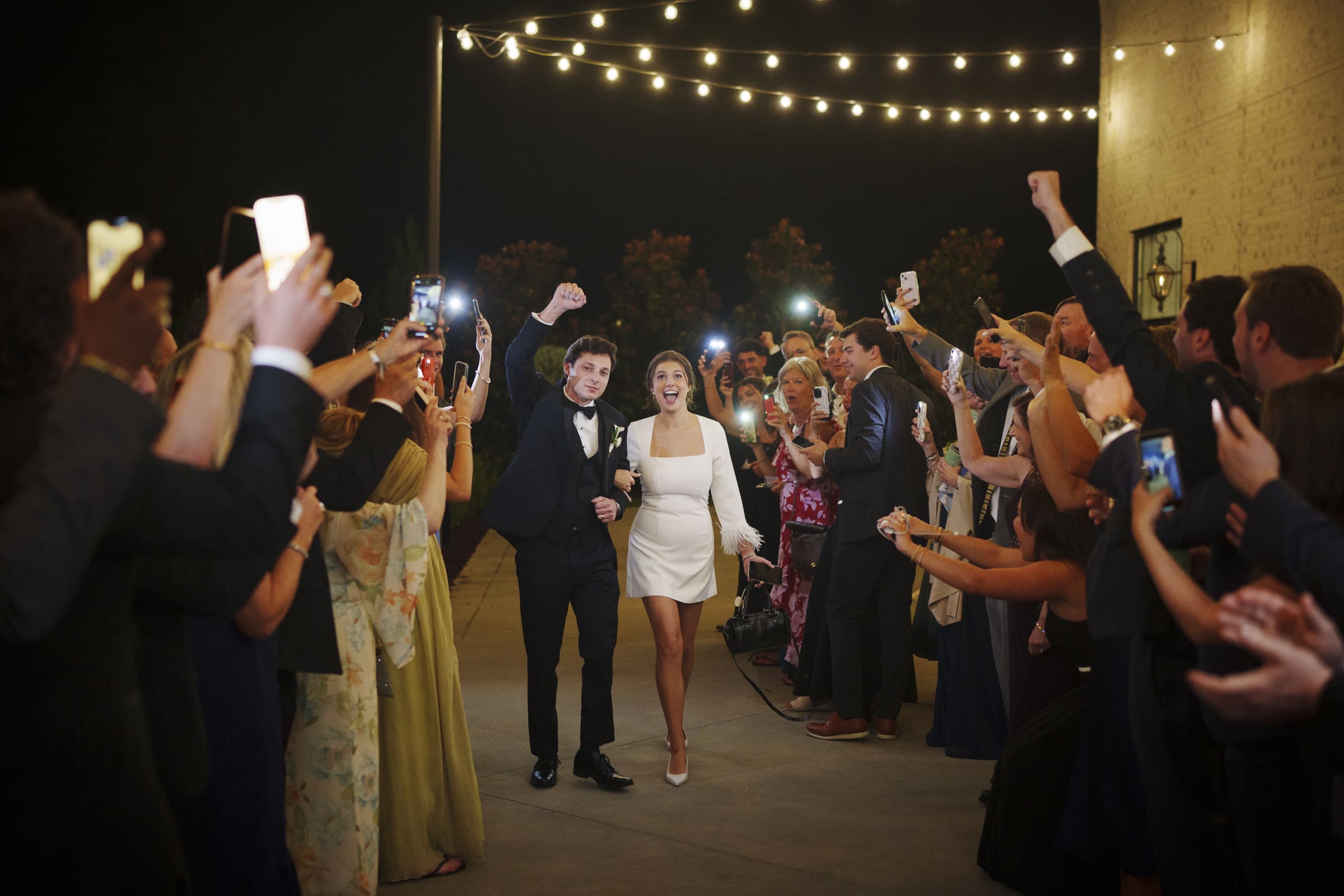 A newlywed couple walks arm-in-arm under string lights at night, smiling as they are cheered on by a crowd of guests holding up phones and raising their arms in celebration.