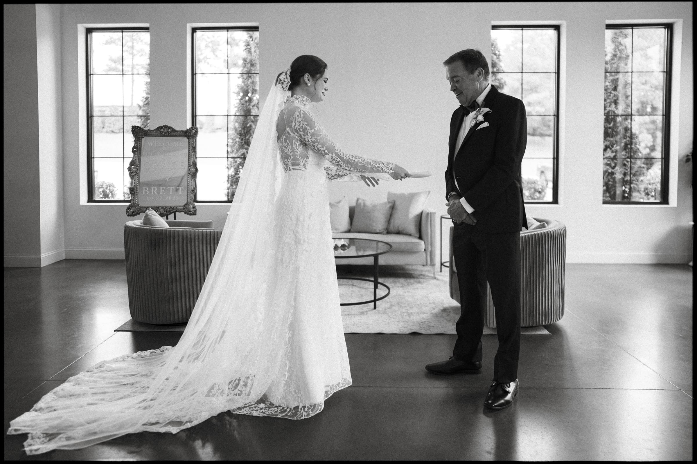 A bride in a long lace gown and veil stands facing an older man in a suit, likely her father, in a bright room with large windows, sofas, and a decorative sign in the background.