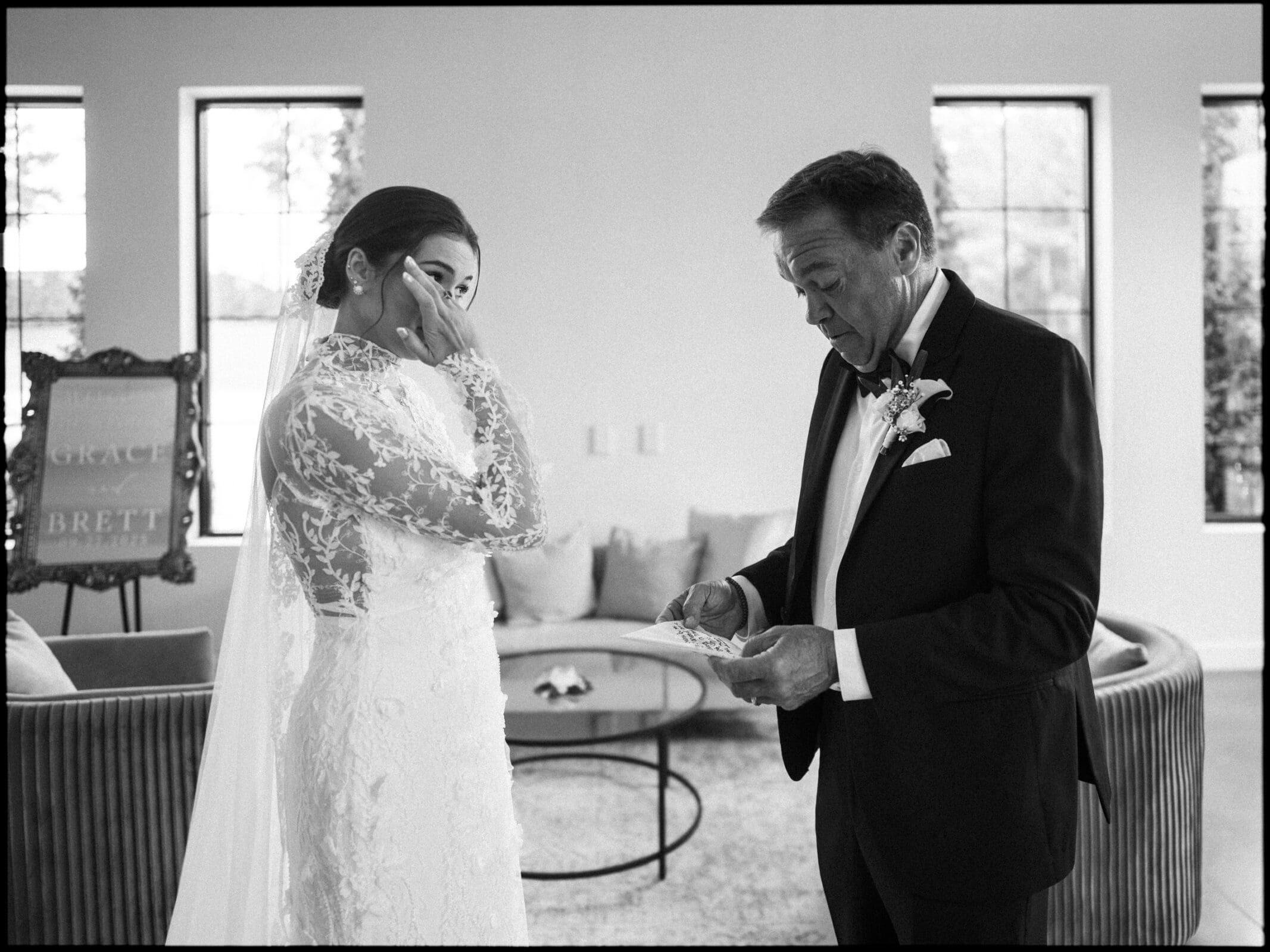 A bride in a lace wedding dress wipes away a tear while standing beside an older man in a tuxedo who is reading a note. They are indoors next to a couch and large windows.