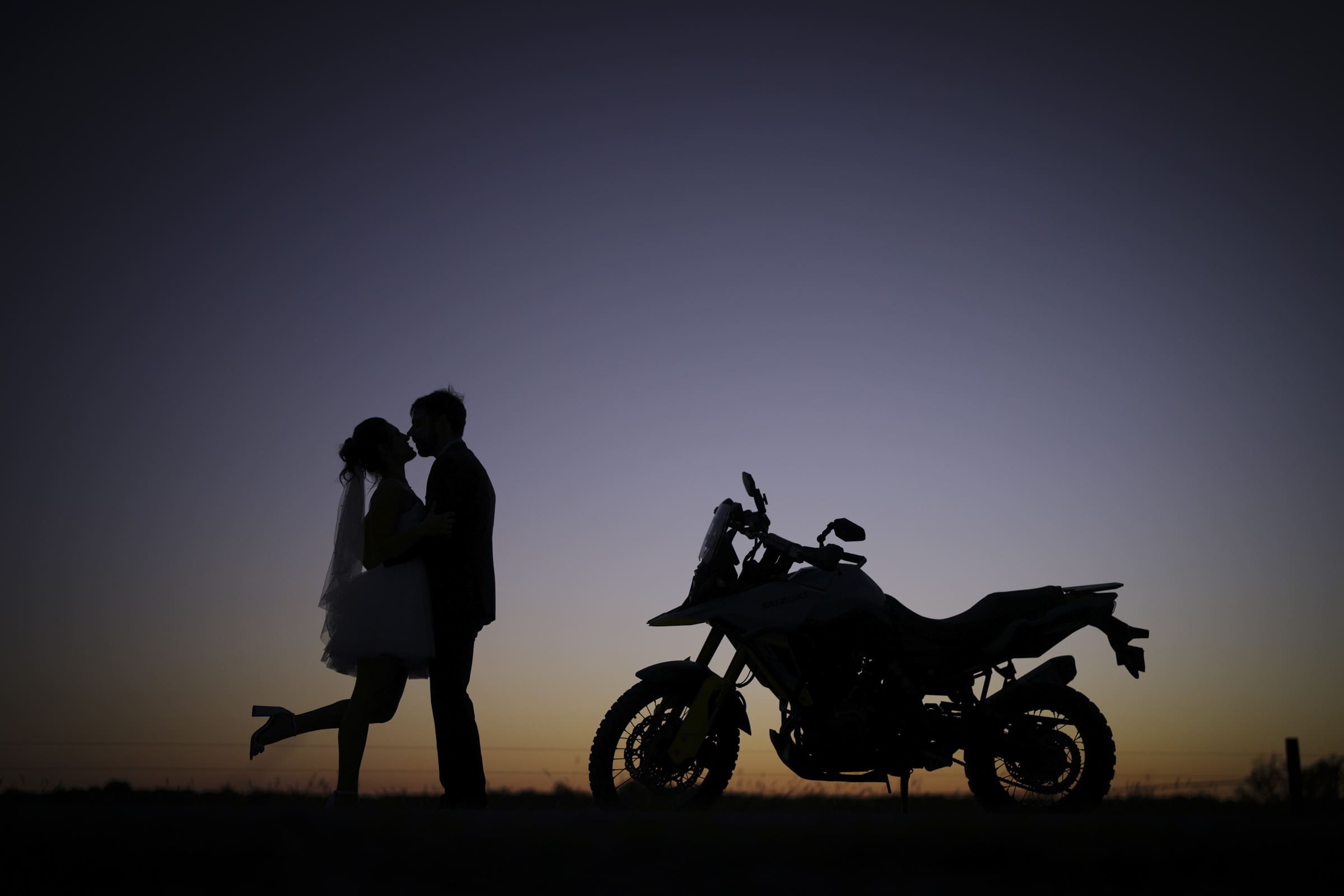 Silhouette of a couple embracing at sunset near a motorcycle; the bride is wearing a veil and lifting one leg while the groom faces her, with a clear sky in the background.