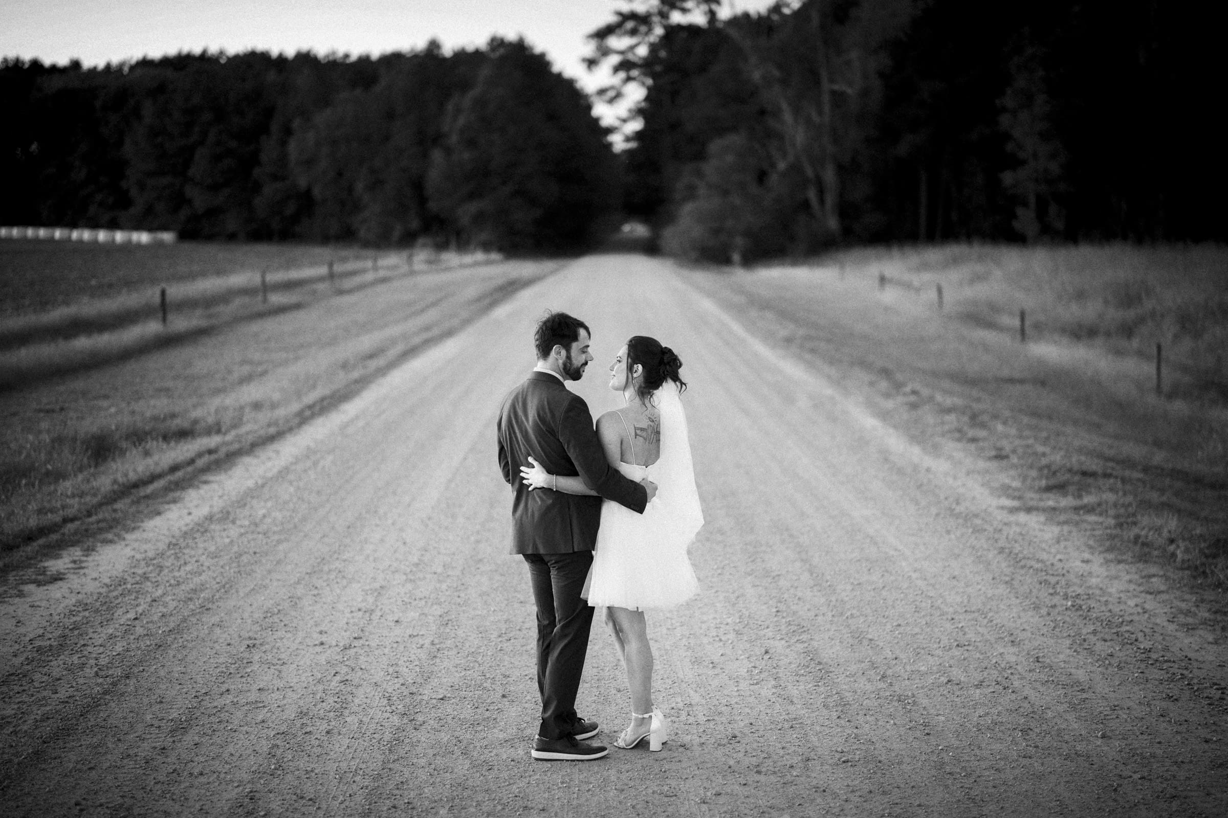 A bride and groom stand on a rural dirt road, holding each other and gazing into each others eyes. Trees and fields line the road, and the scene is in black and white.