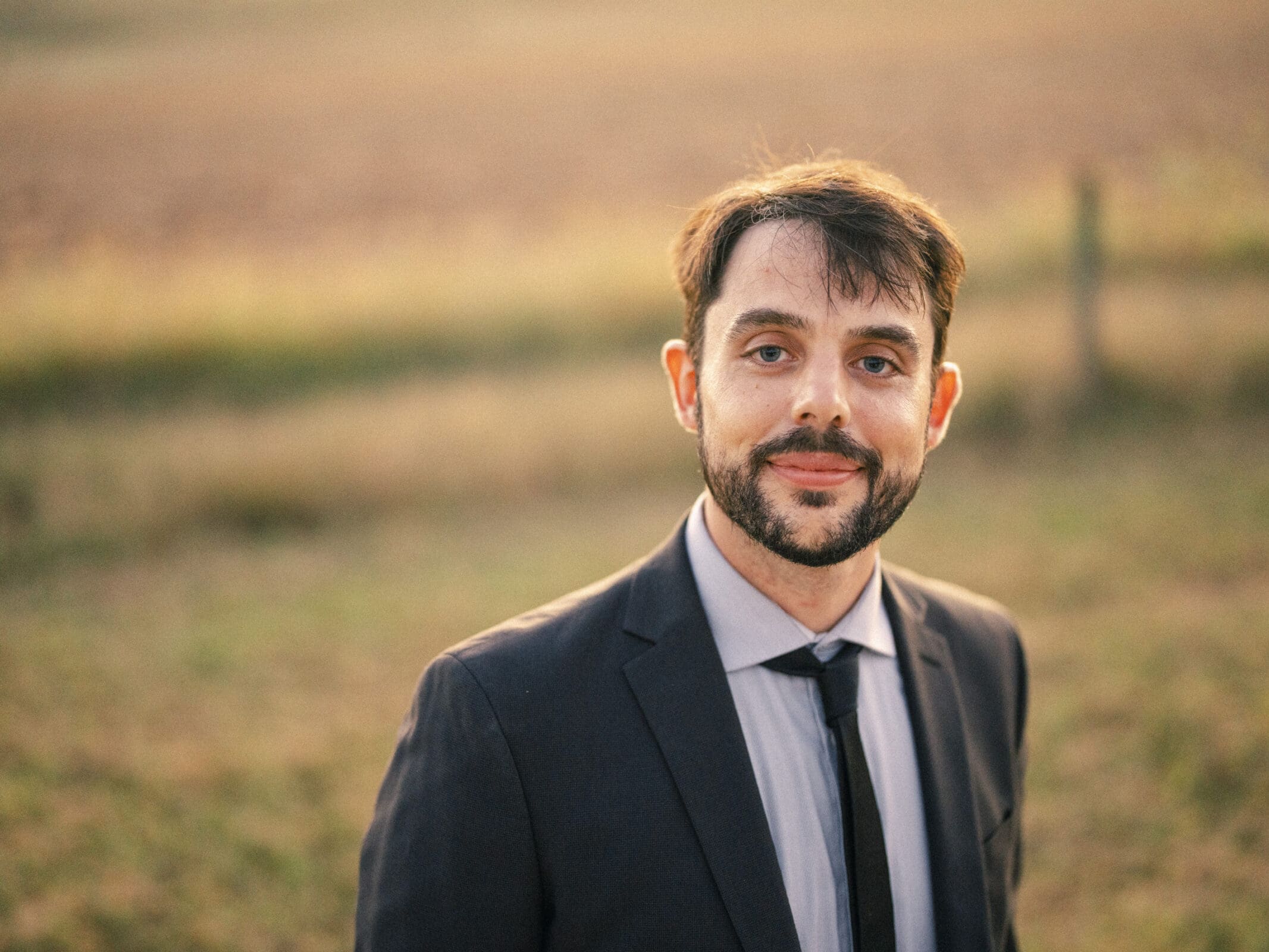 A man in a suit and tie stands outdoors in a grassy field at sunset, smiling slightly at the camera. The background is softly blurred with warm, golden light.