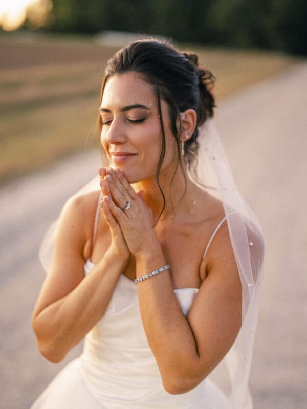 A bride in a white dress and veil stands outdoors on a sunlit path, eyes closed and hands clasped near her face, appearing peaceful and content.