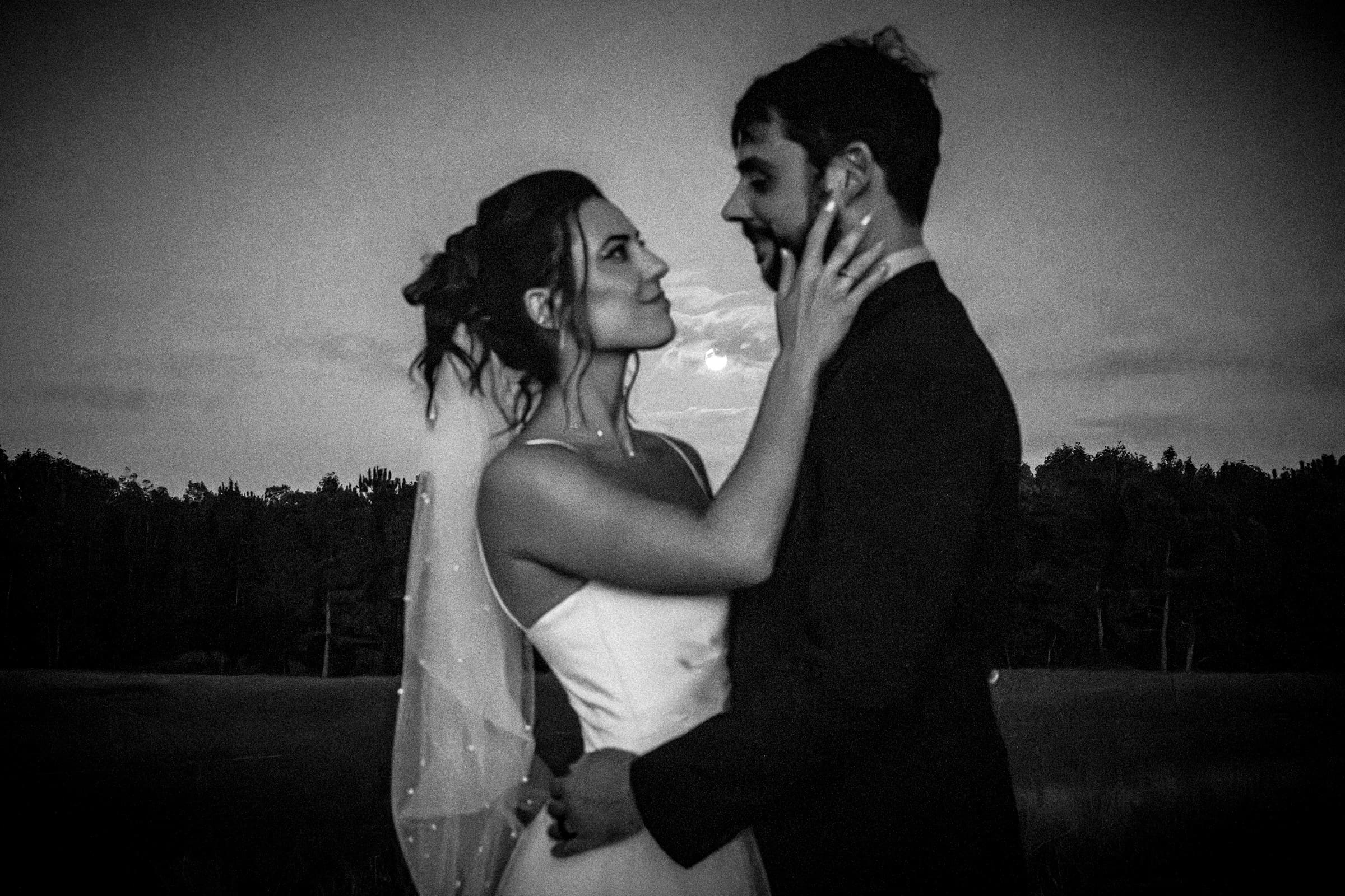 A bride and groom stand closely, gazing into each others eyes and holding each other tenderly outdoors at dusk, with trees in the background. The image is in black and white, creating a romantic, timeless feel.