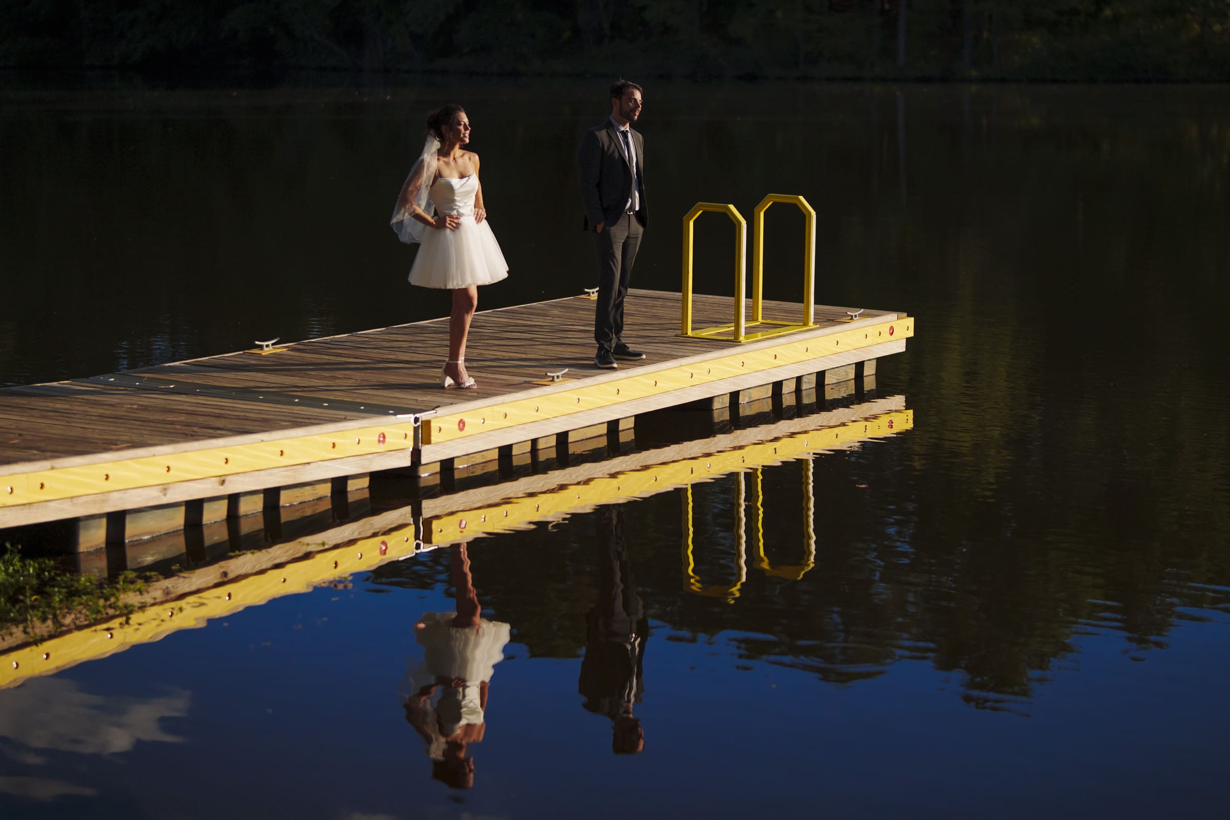 A bride in a short white dress and a groom in a suit stand on a wooden dock over calm water, their reflections visible in the lake below them. The scene is bright and serene, surrounded by nature.