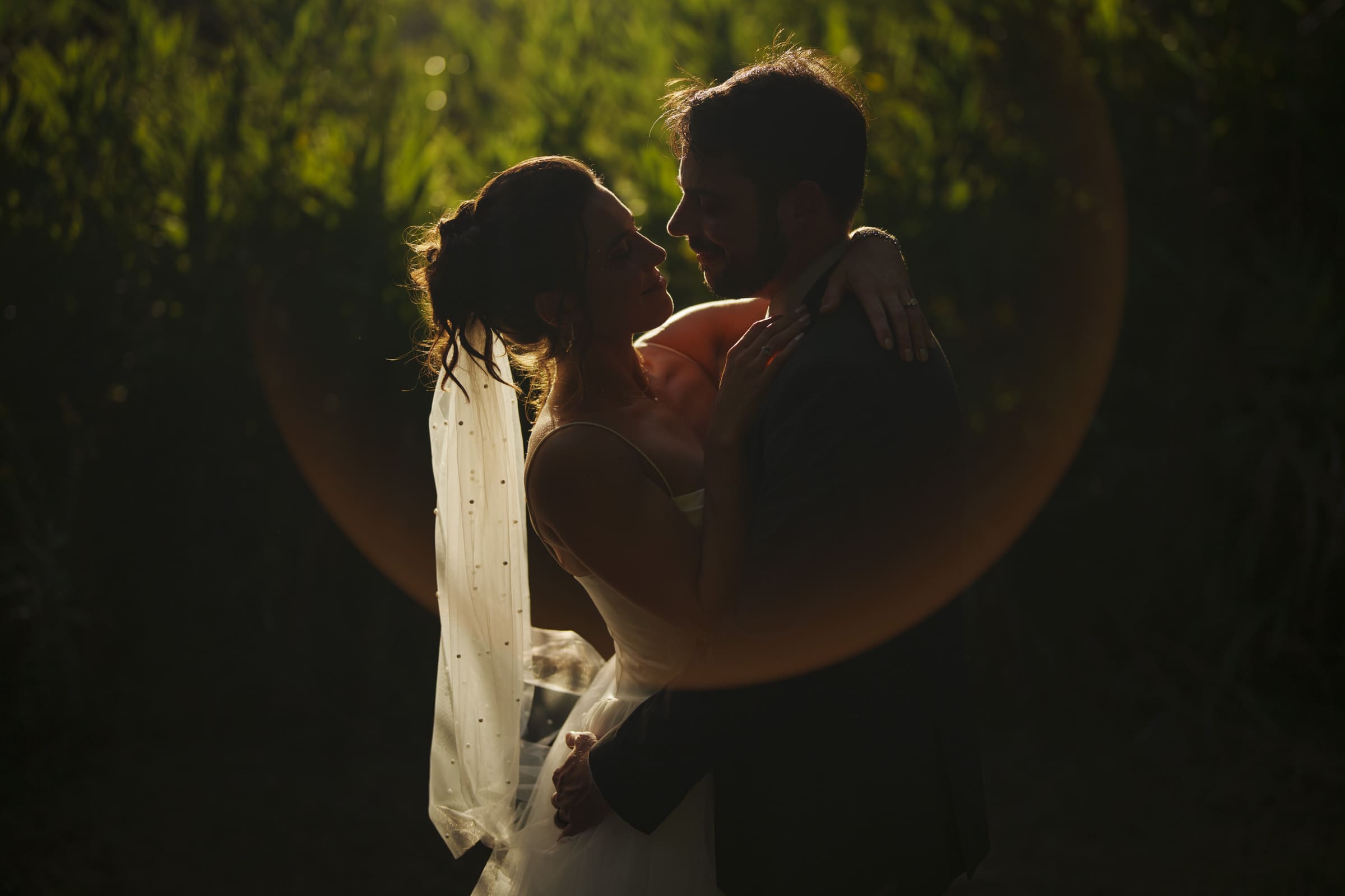 A bride and groom embrace outdoors in soft, golden sunlight, surrounded by greenery. The bride wears a white dress and veil, and both look lovingly at each other, framed by a subtle halo of light.