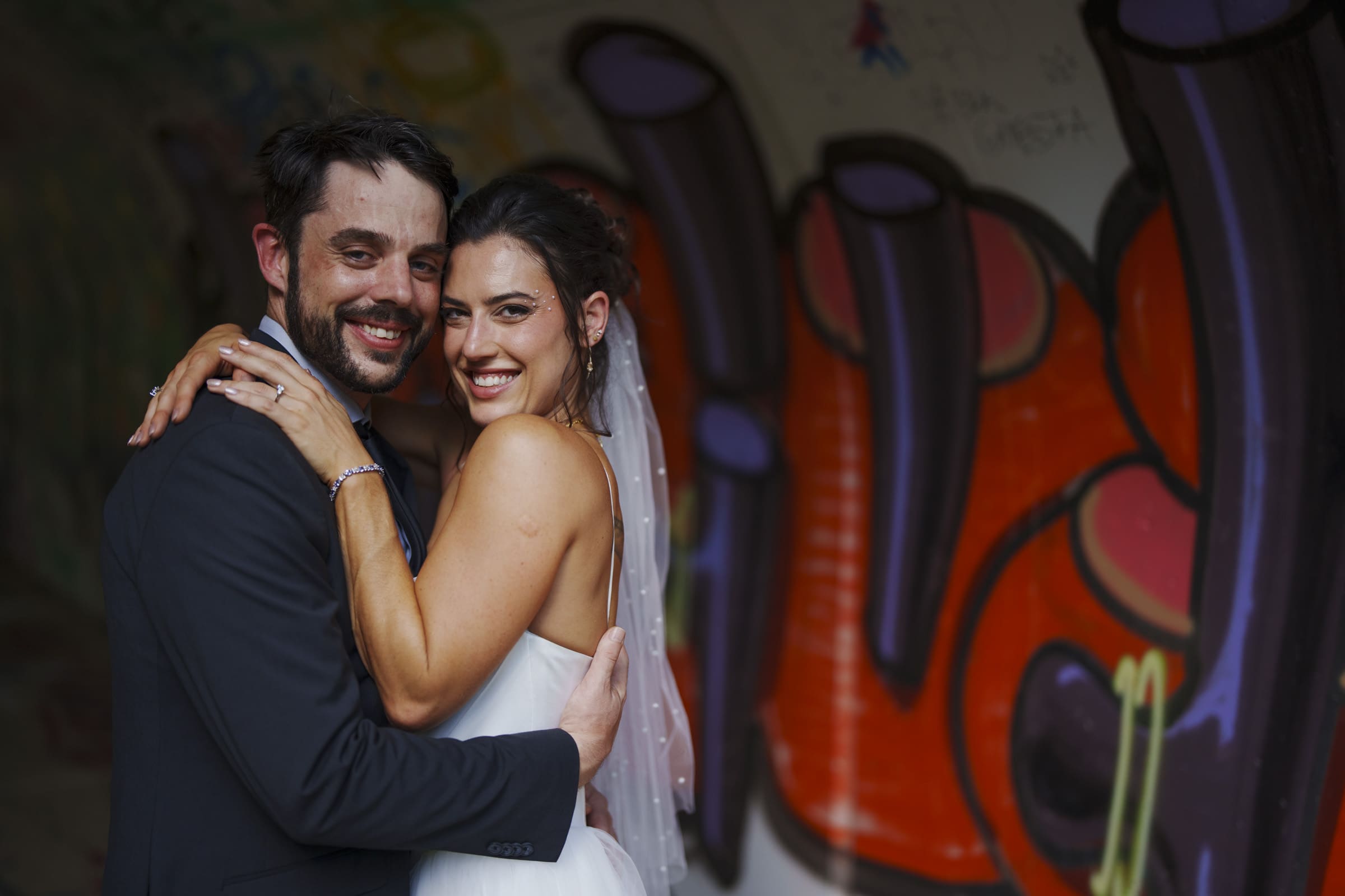 A bride and groom dressed in wedding attire smile and embrace in front of a colorful wall covered in graffiti art.