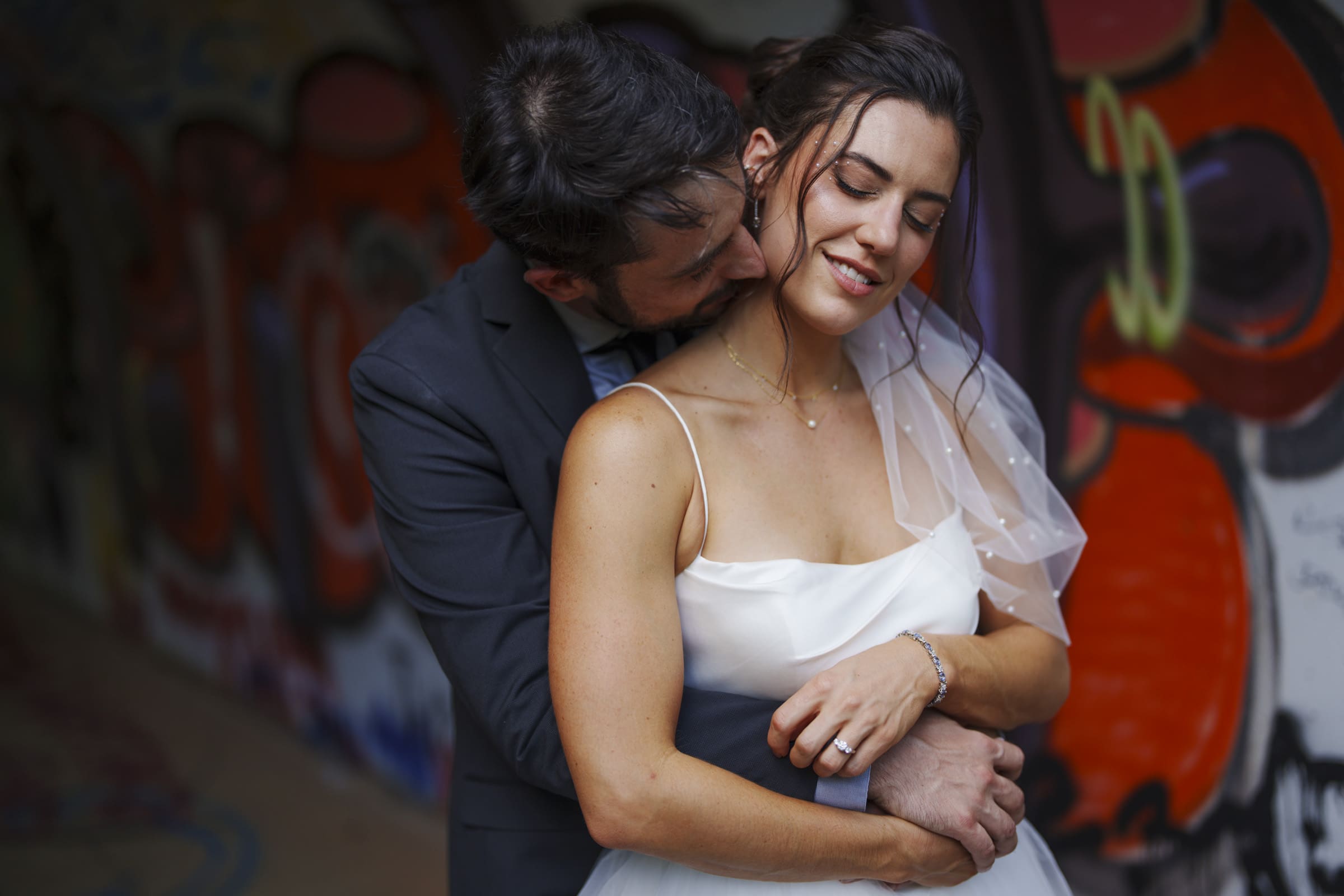 A groom lovingly embraces his bride from behind, nuzzling her neck. The bride, dressed in a white gown and veil, smiles softly with closed eyes. Colorful graffiti is visible in the blurred background.
