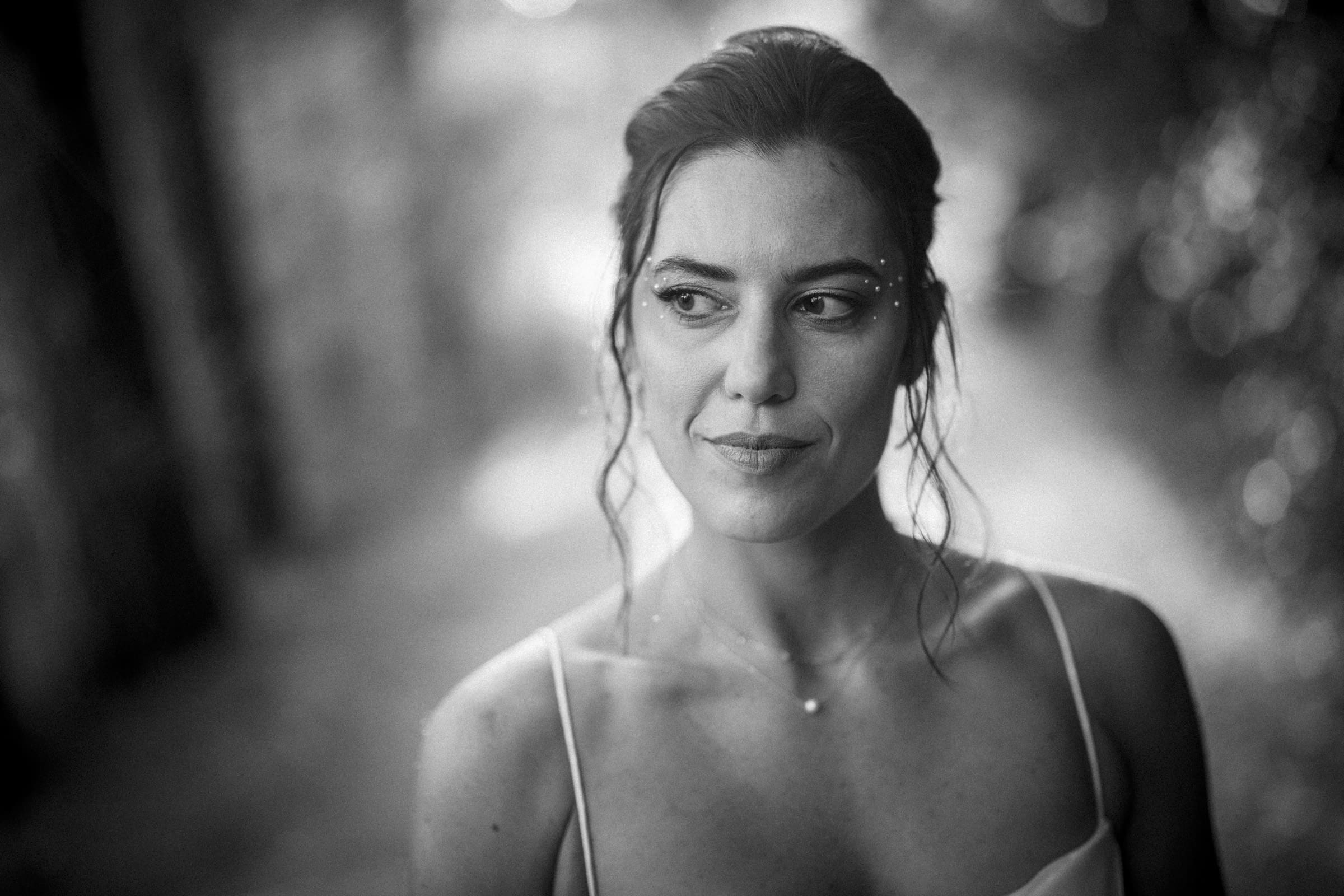A woman with loose hair strands and subtle makeup, including rhinestones near her eyes, stands outdoors in soft focus light, wearing a thin-strapped top, and looks pensively to the side. The photo is in black and white.