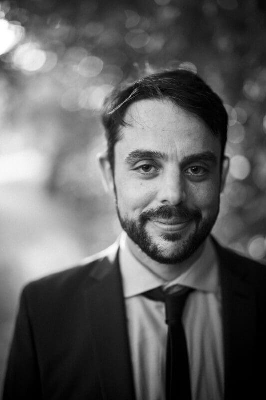 A man with short dark hair, a beard, and a mustache is wearing a suit and tie, smiling softly at the camera. The background is blurred with circular light spots, creating a bokeh effect.