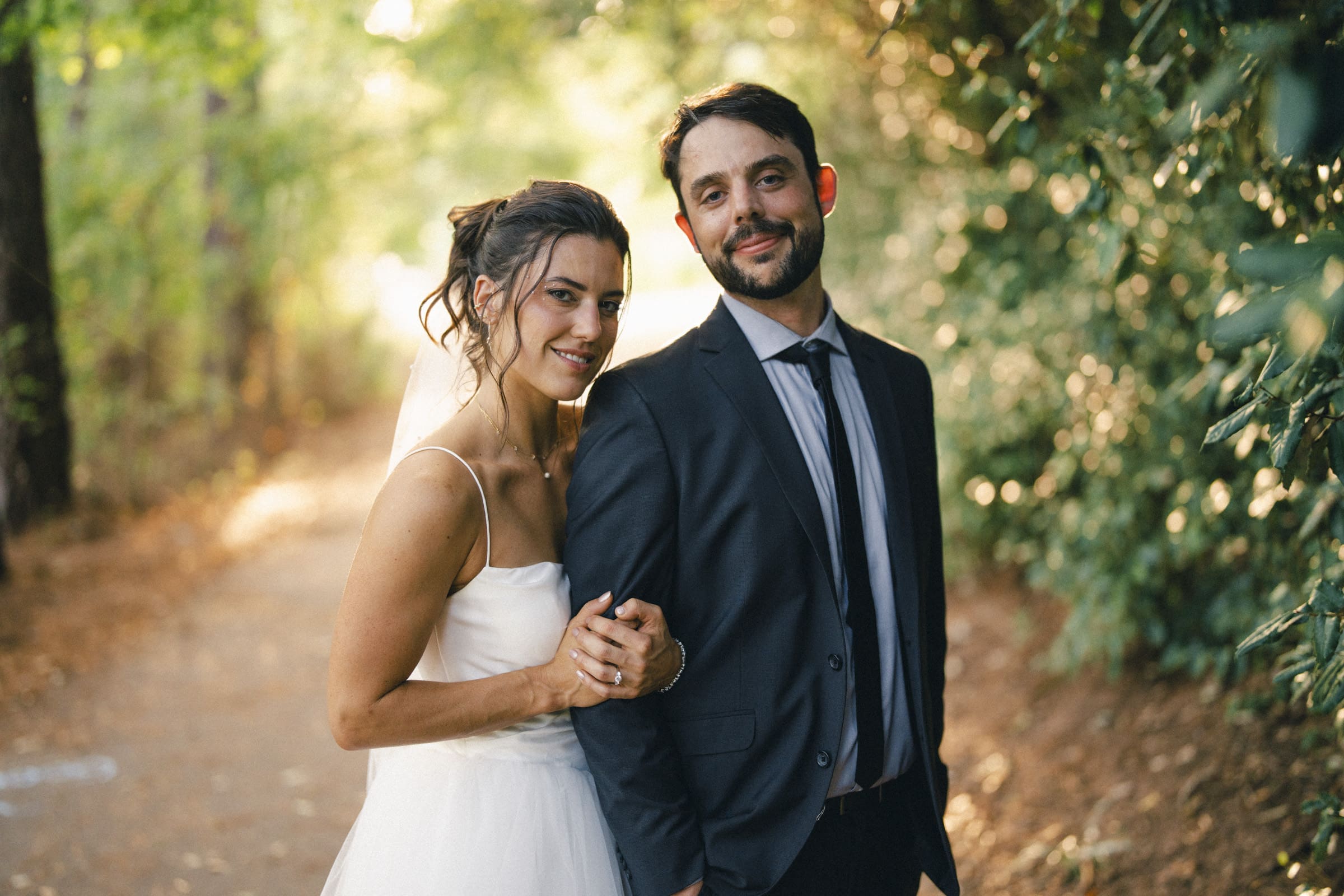 A bride in a white dress and veil stands closely beside a groom in a dark suit and tie, smiling together on a sunlit wooded path surrounded by green foliage.