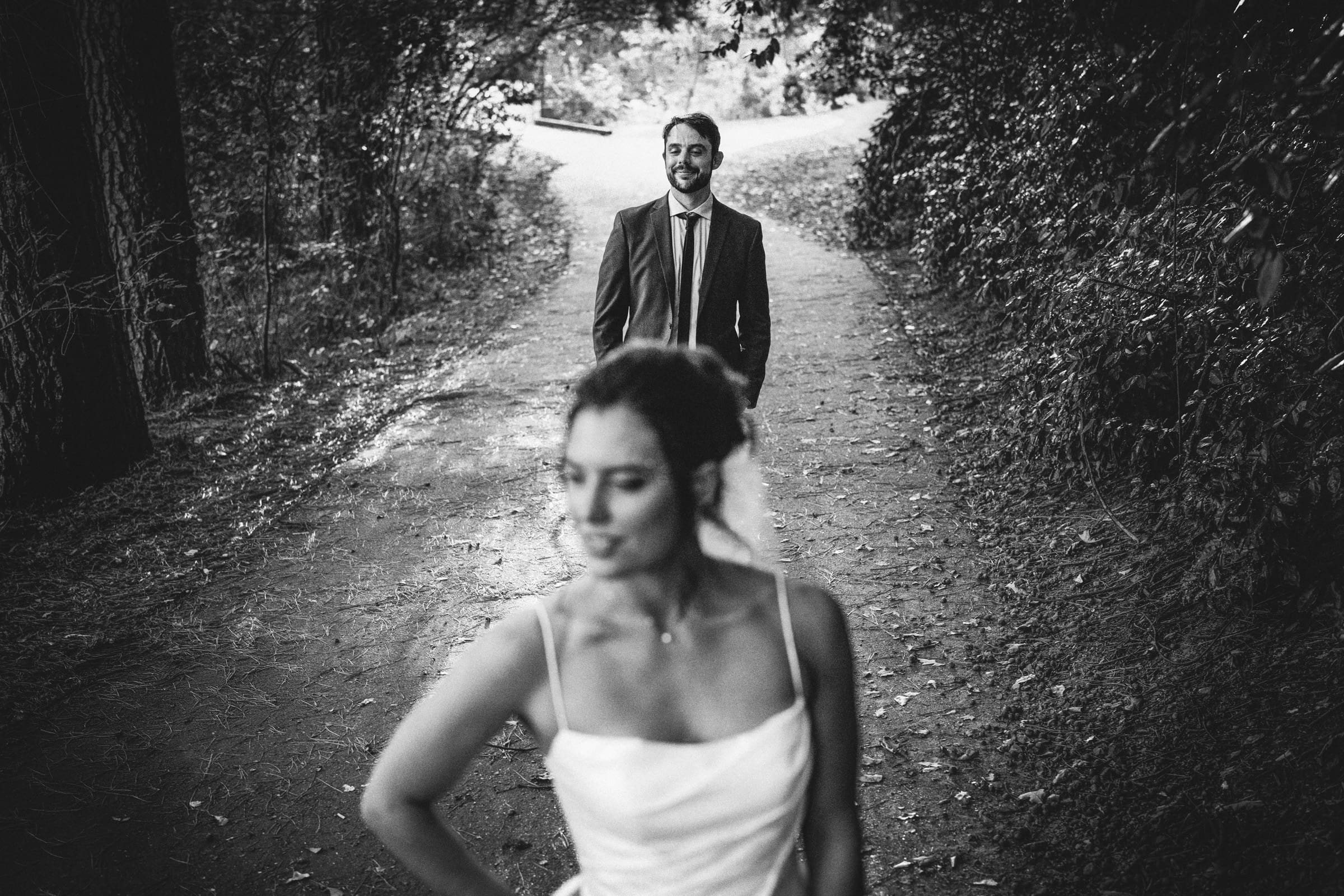 A black-and-white photo of a bride standing in the foreground with her back to a groom, who is smiling and standing on a tree-lined path in the background.