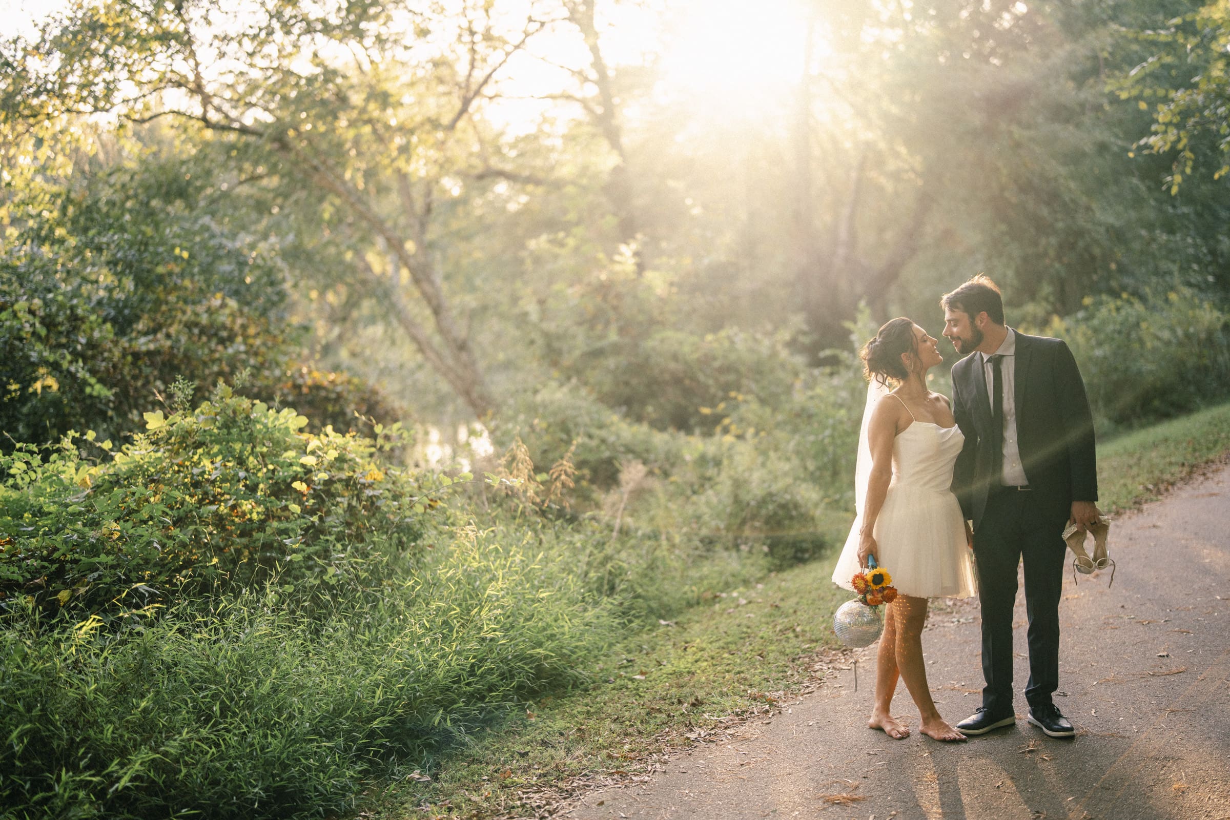 A bride and groom stand barefoot on a sunlit path in a lush, green park. The bride holds a bouquet and the groom holds his shoes. They smile at each other as sunlight streams through the trees around them.