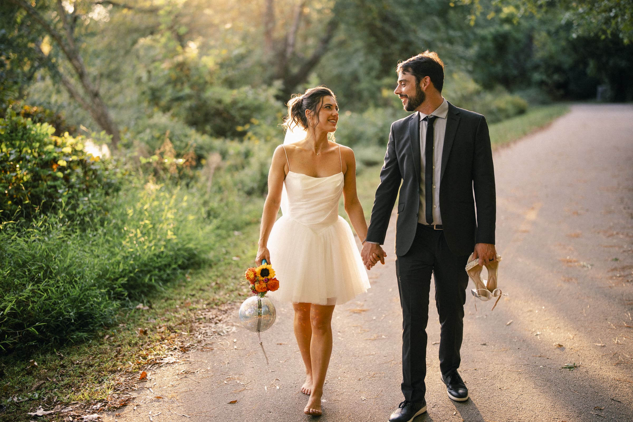 A barefoot bride in a short white dress holds flowers and a balloon, walking hand-in-hand with a man in a dark suit on a sunlit path surrounded by green trees, both smiling at each other.