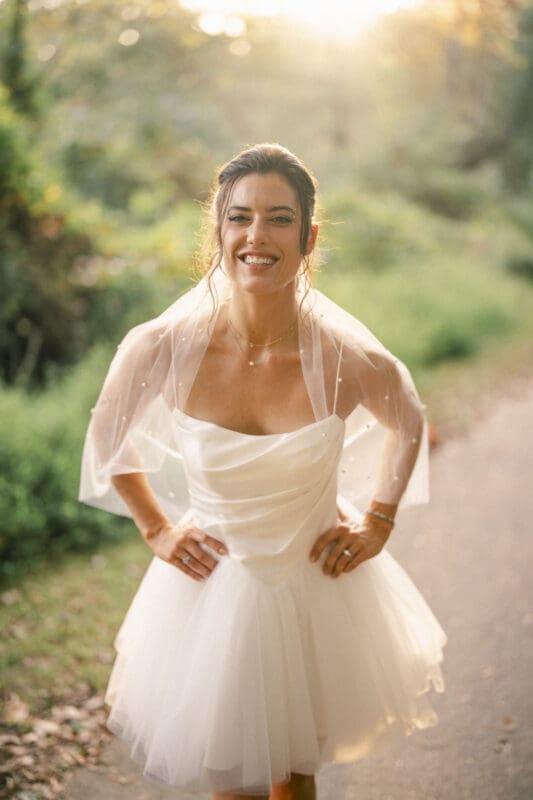 A smiling bride in a short white wedding dress and sheer veil stands outdoors on a sunlit path, surrounded by greenery, with her hands on her hips.