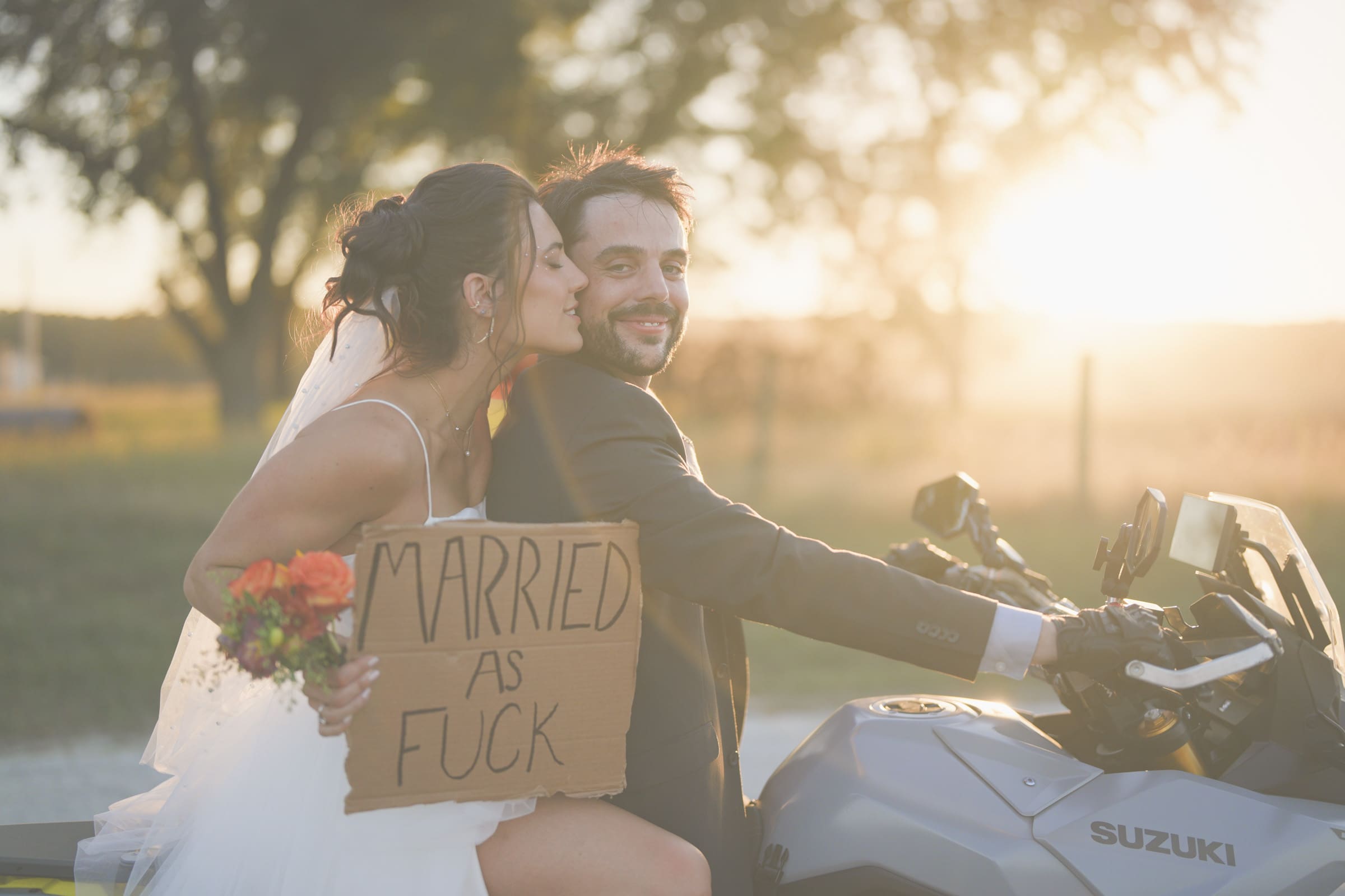A bride and groom sit on a motorcycle at sunset. The bride holds a bouquet and a cardboard sign that says MARRIED AS FUCK, while kissing the groom on the cheek. Both appear happy and relaxed.
